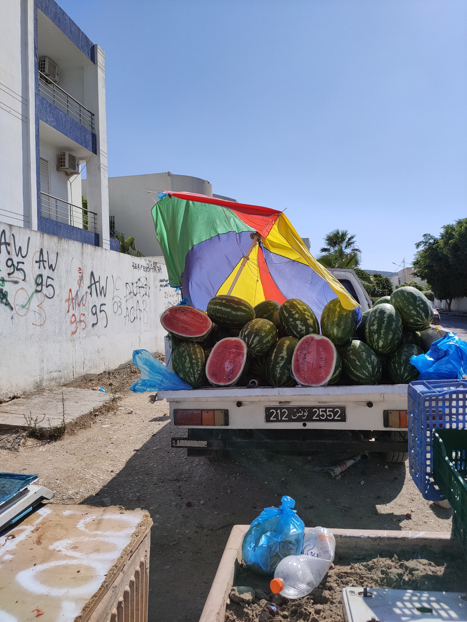An outdoor medium shot captures a white pickup truck loaded with watermelons, parked on a dirt and gravel surface in front of a white and blue building. The truck is adorned with a colorful umbrella, predominantly red, yellow, and blue, providing shade over the fruit. Several watermelons are visible, with some cut in half, revealing their red flesh. Blue plastic bags and a blue crate are also present on or near the truck. The building behind the truck has multiple stories with balconies and air conditioning units. Graffiti adorns the wall of the building on the left. The ground is uneven, with scattered debris. A portion of a weighing scale and a wooden crate are in the foreground on the left. Several plastic bottles and a blue trash bag are in a small concrete enclosure in the lower right corner. The license plate on the back of the truck reads "212 2552" in both Arabic and Latin characters. The scene appears to be a roadside market or vendor.