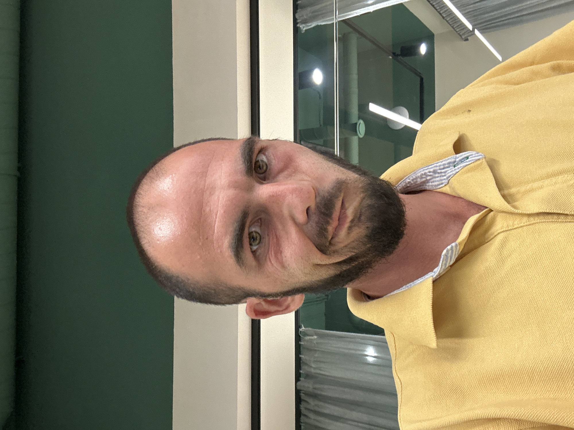 The image is a head-and-shoulders selfie of a man with fair skin, a receding hairline, and a dark beard. He is wearing a yellow polo shirt with a striped collar. His facial expression is a smirk, with his mouth slightly twisted to one side. The background appears to be an indoor setting, possibly an office or commercial space, with a dark green ceiling, white walls, and modern lighting fixtures visible. There are also sheer curtains and windows in the background, suggesting the presence of natural light or a view to the outside. The man's gaze is directed towards the camera. The scene has no discernible markers indicating a specific location within Budva, Montenegro.