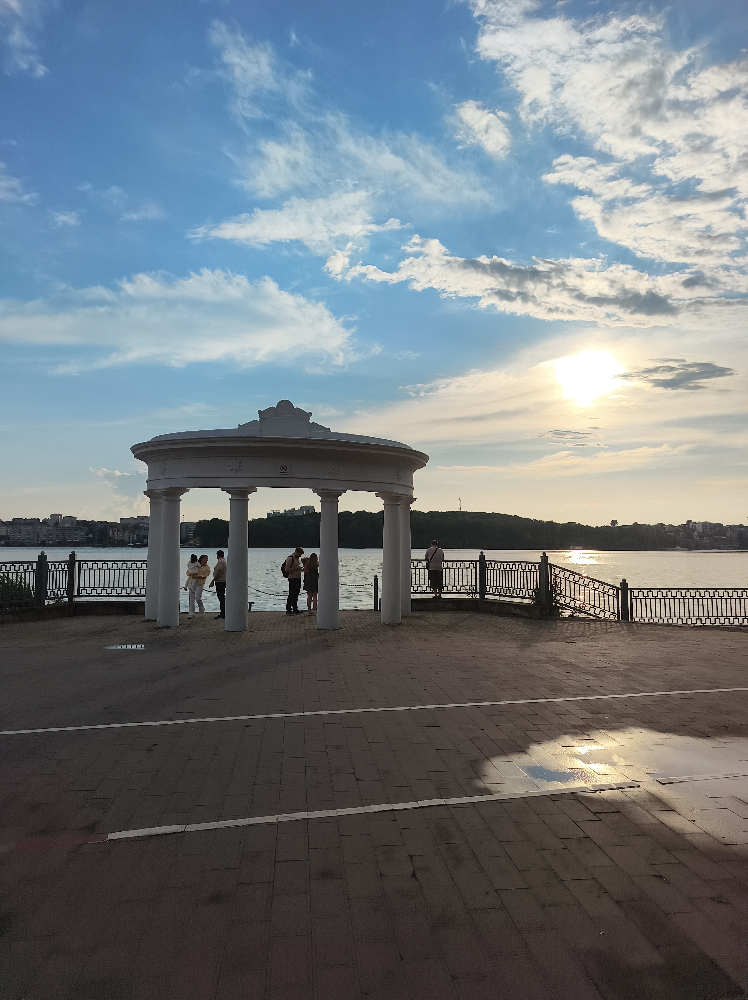 The image depicts a waterfront promenade in Ternopil, Ukraine, on a cloudy day with the sun partially visible. A white rotunda with classical columns stands on the promenade, overlooking a body of water. Several people are present: a couple with a child stand near the rotunda, and another two individuals are conversing within its structure. Further along the promenade, another person stands by the railing, looking out at the water. The promenade is paved with stone tiles, and a metal fence lines the water's edge. In the distance, a city skyline with buildings and trees is visible across the water, with a treed hill forming the background. The sky is a mix of blue and white clouds, with the sun casting a glare on the water.