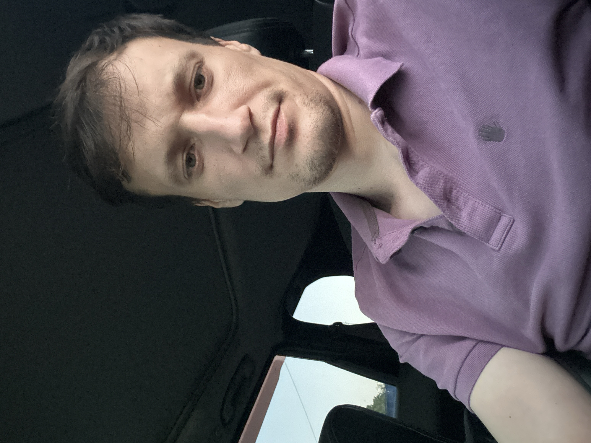 A selfie shows a light-skinned male with brown hair and light brown eyes sitting in the driver's seat of a car. He is wearing a purple polo shirt with a small grey hand logo on the left breast. His expression is neutral, and his gaze is directed towards the camera. The car's interior is visible, including the black headliner and a sunroof. The background outside the car windows shows a pale blue sky and some greenery. The scene is set during daylight hours.