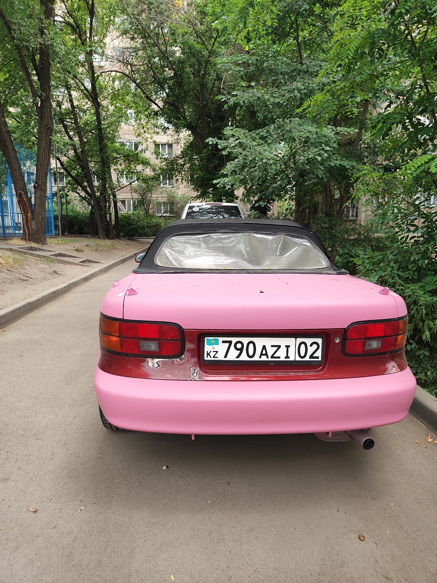 A pink and red convertible car is parked on a paved surface in Almaty, Kazakhstan. The car has a black convertible top that is covered with clear plastic sheeting. The license plate on the back of the car reads "KZ 790 AZI 02". The car appears to be parked on the side of a street or in a parking lot, with trees and a building visible in the background. The building is a multi-story structure with visible windows and a light-colored facade. The environment is characterized by lush green foliage from trees and bushes. The ground is a light gray paved surface, and there is a concrete curb separating the paved area from a sandy strip of ground. There are no people visible in the image.