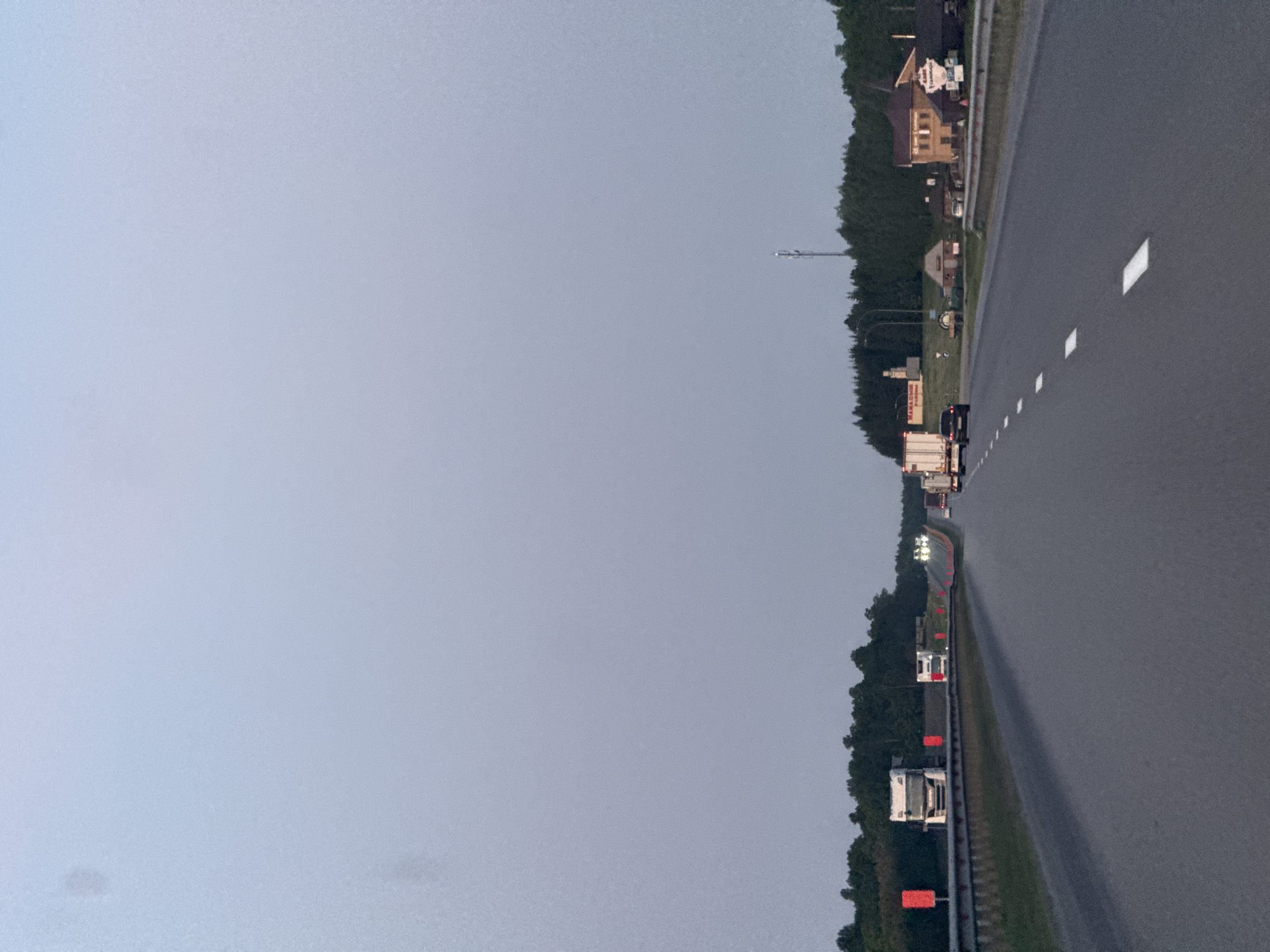 The image depicts a roadway in Bol'shoi Mashlyak, Russia, under a cloudy sky. The roadway is lined with trees and roadside objects and is marked with dashed lane lines. Several trucks and cars are traveling along the roadway. In the distance, buildings are visible.