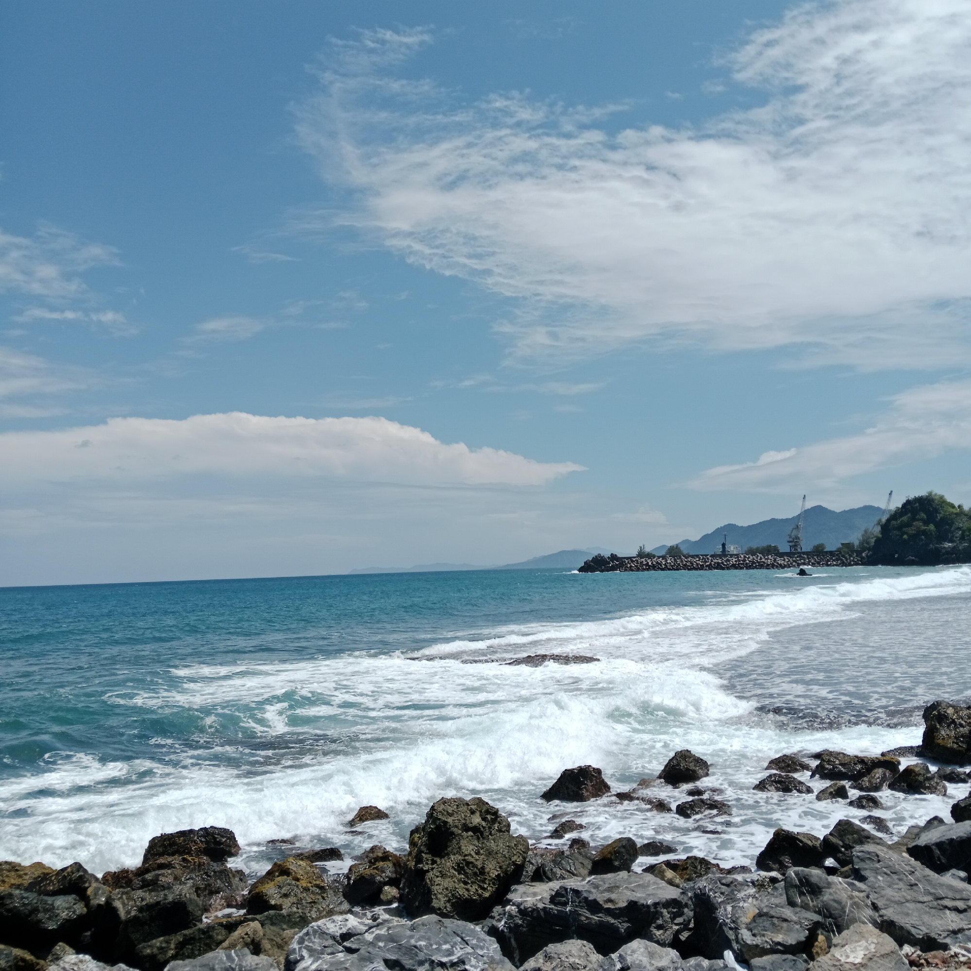 The image depicts a rocky shoreline on a bright, clear day. The foreground is dominated by a collection of dark, weathered rocks of varying sizes, some partially submerged in the water. The ocean is a vibrant turquoise blue, with white waves crashing onto the shore, creating a foamy surf. In the mid-ground, a long breakwater made of large rocks extends into the sea, with industrial cranes visible on the land beyond. The background features a range of mountains covered in lush green vegetation, under a bright blue sky dotted with wispy white clouds. The scene is devoid of people. This location is identified as Naga Umbang, Indonesia.
