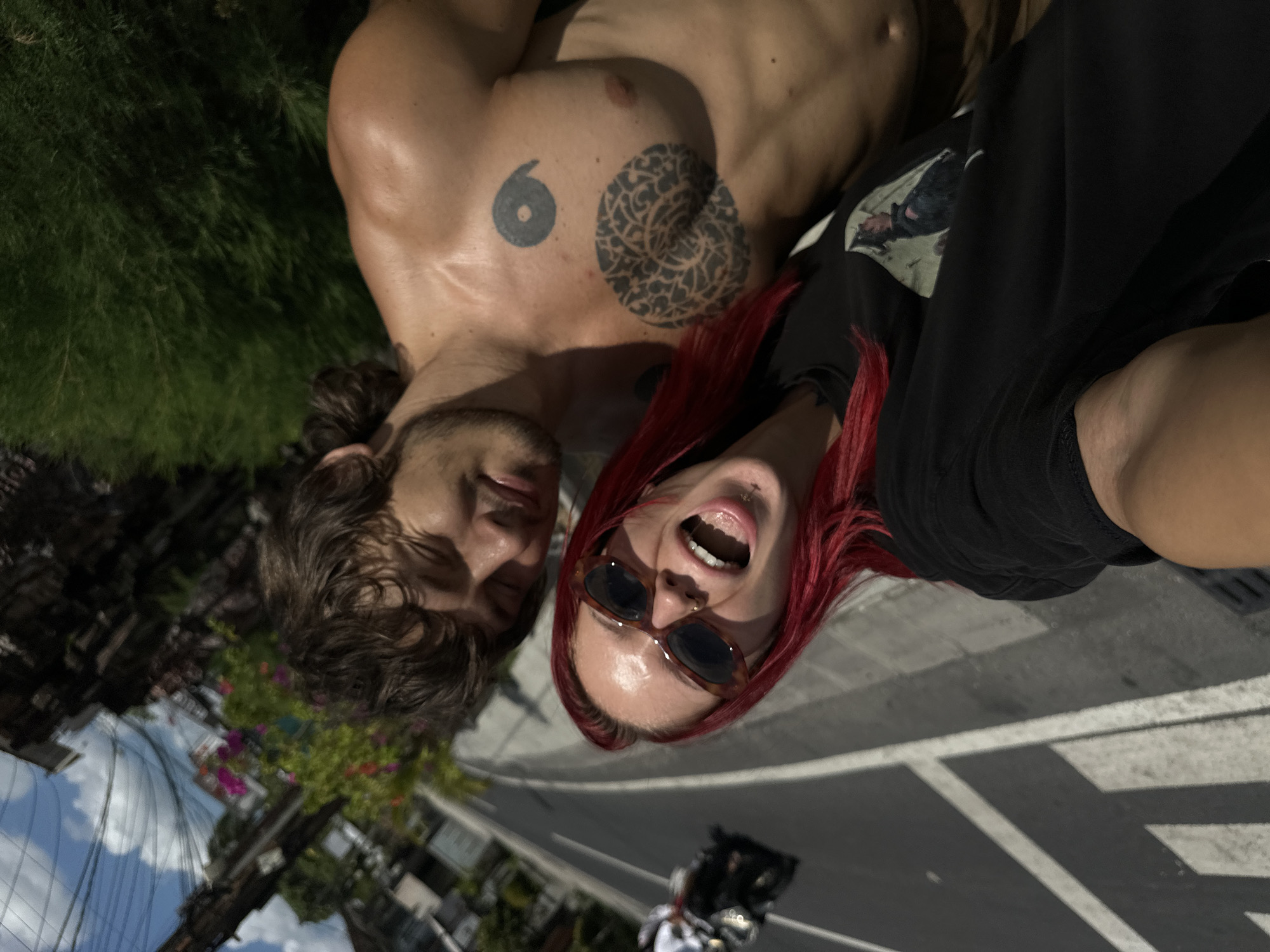 A selfie photograph depicts two individuals: a shirtless man with dark hair and a woman with red hair, both facing the camera from a low angle on the street in North Kuta, Indonesia. The man, positioned on the left, has several tattoos, including a number 6 and a circular design on his chest. The woman wears sunglasses and a black t-shirt, with her mouth open in a possible expression of excitement. The background features a paved road, with a blurred image of a moped and driver. Vegetation and buildings are visible in the periphery. The sky is visible in the reflection on the ground, indicating a bright, daytime setting.