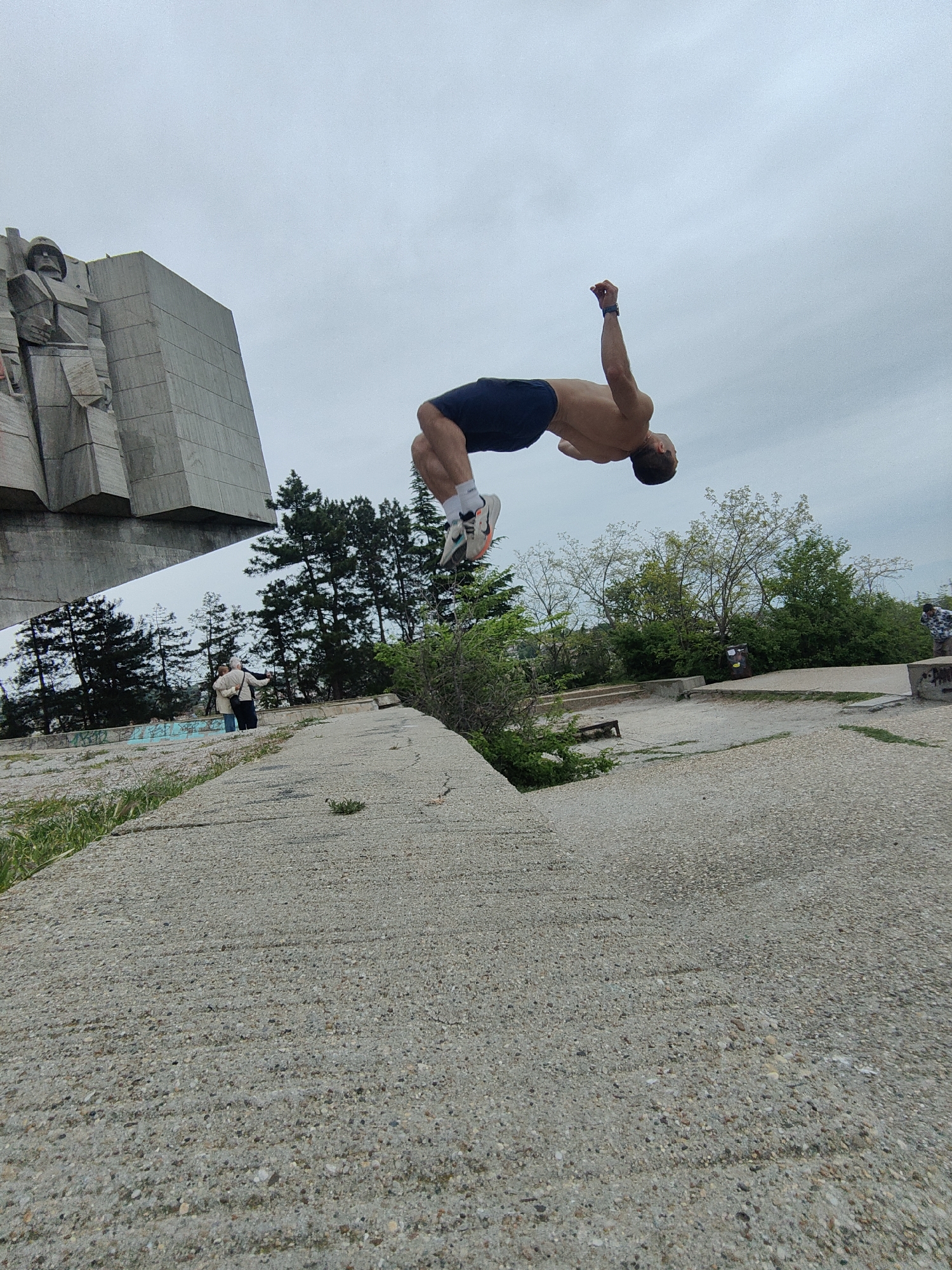 A man is captured mid-backflip in an outdoor location, seemingly a park or public space within the city of Varna, Bulgaria. He is shirtless, wearing dark shorts and white socks, and athletic shoes. His body is arched backward with his arms extended. To the left stands a large concrete structure, partially visible, appearing to be a segment of a monument or sculpture, likely associated with a communist-era memorial. Two people are visible in the background standing near the monument with the trees. The terrain is uneven concrete with grass. The sky is overcast.