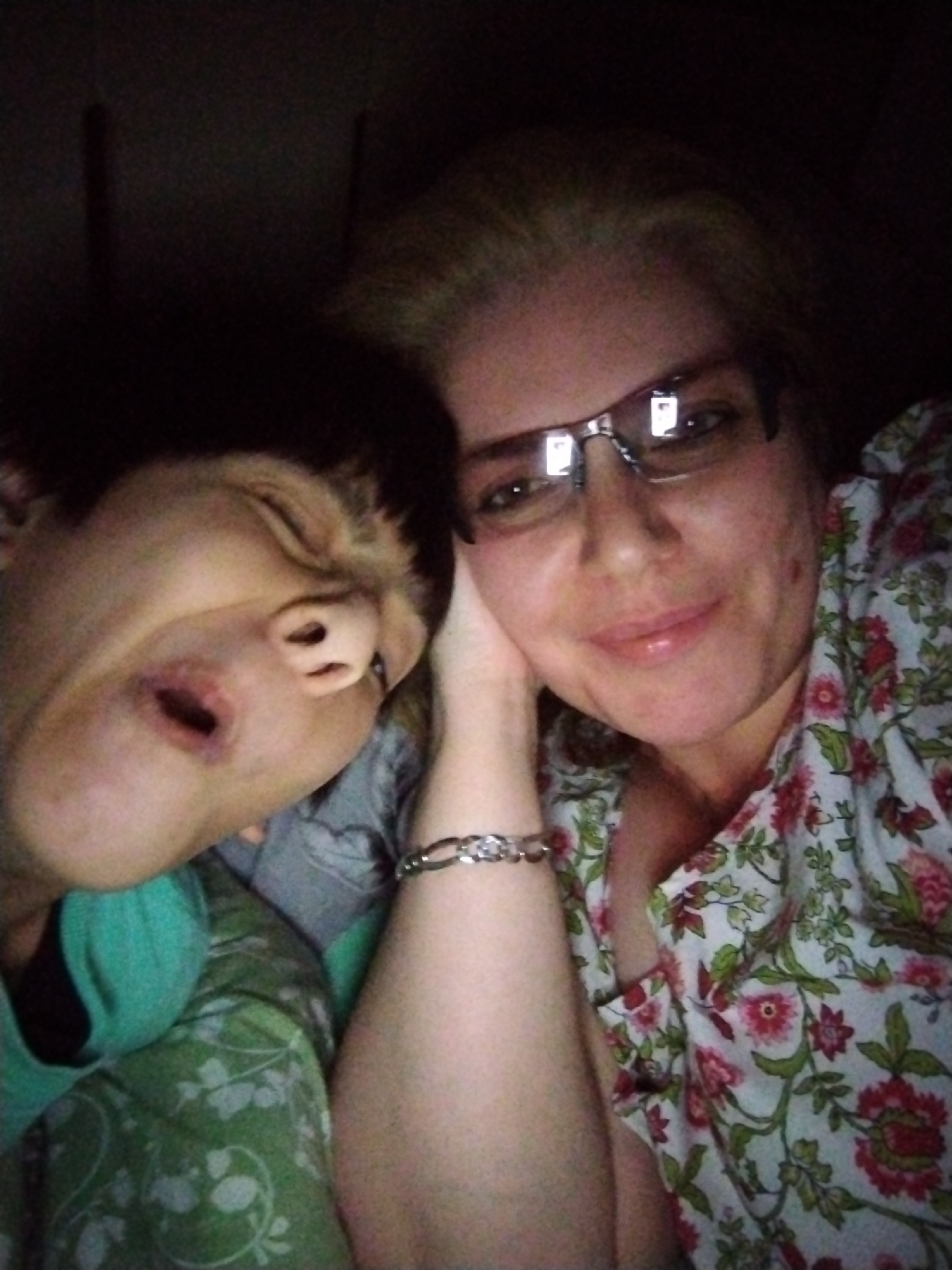 The image displays a close-up, dimly lit portrait of two individuals in an indoor setting. On the right, an adult female with blonde hair, wearing rectangular glasses, is seen smiling and looking directly at the camera. She wears a light-colored top with a prominent red, pink, and green floral pattern. Her left arm is bent, with her hand supporting her head or cheek, and a silver chain-link bracelet is visible on her left wrist. Reflections of an artificial light source are apparent in her eyeglass lenses. To the left, partially obscured, is a younger individual with dark hair, appearing to be asleep or resting, with eyes closed and mouth slightly open. This individual is wearing a green top, with a green and white patterned fabric, possibly a blanket or another garment, visible beneath their head. The two subjects are positioned intimately close to each other. The background is dark and largely undifferentiated, with no discernible objects or features. There is no visual information within the image itself to indicate the location as the city of Al Mas'oudi, United Arab Emirates, or any other specific geographical area.
