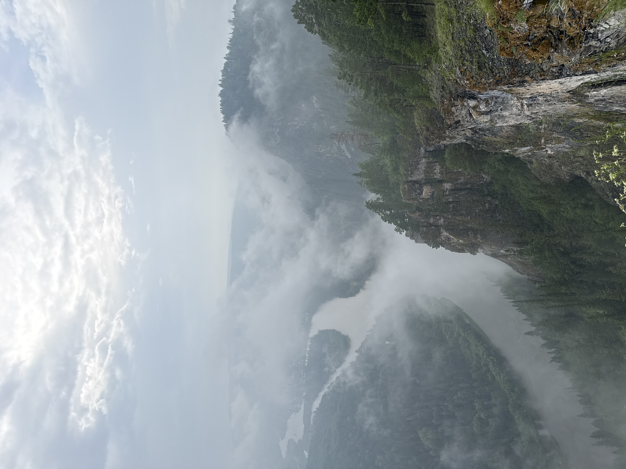 A high-angle, medium shot captures a scenic view from a cliffside overlooking a misty river valley. The foreground features a rocky cliff face with sparse vegetation and some dried grasses. Dense evergreen trees cover the slopes leading down to the river. The river itself winds through the valley, partially obscured by swirling fog and mist. On the far side of the river, more forested hills are visible, also shrouded in mist, with a few structures suggesting a small settlement. The sky above is overcast with a soft, diffused light filtering through the clouds. The overall atmosphere is serene and somewhat mysterious due to the fog. The scene is located in the vicinity of Urman, Russia, known for its natural beauty and river landscapes. There are no people or animals visible in the image.