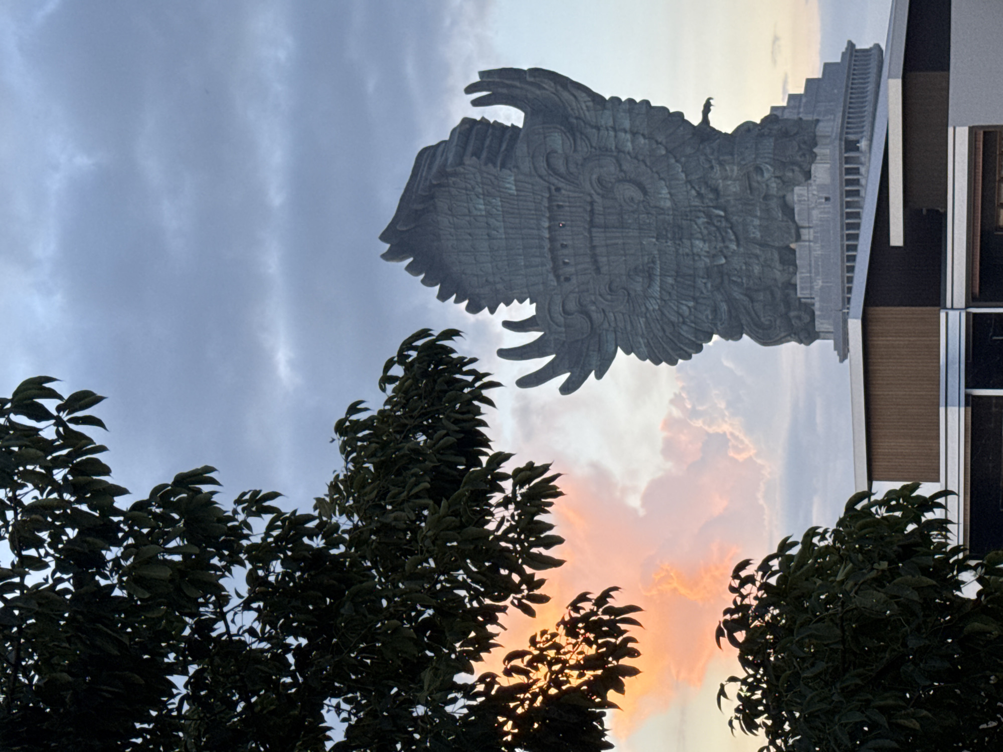 The image, presented rotated 90 degrees counter-clockwise, depicts a monumental statue of a mythical bird or Garuda set against a vibrant sky, likely at sunset or sunrise. The colossal sculpture, featuring intricate details on its head and powerful, textured wings, occupies the upper half of the frame. A tiny human figure is visible standing on top of the statue's head, emphasizing its immense scale. This is likely the Garuda Wisnu Kencana statue in Kuta Selatan, Indonesia.
Behind the statue and extending to the right, a multi-tiered building or base structure is visible, integrated with the artwork. The sky in the background displays a blend of soft grey-blue clouds and warmer, glowing orange-pink hues, particularly towards the lower right, indicative of either late afternoon or early morning light. Dark, silhouetted foliage from trees frames the lower and left portions of the image, with some leaves subtly illuminated by the warm light from behind. No text is visible in the media file.