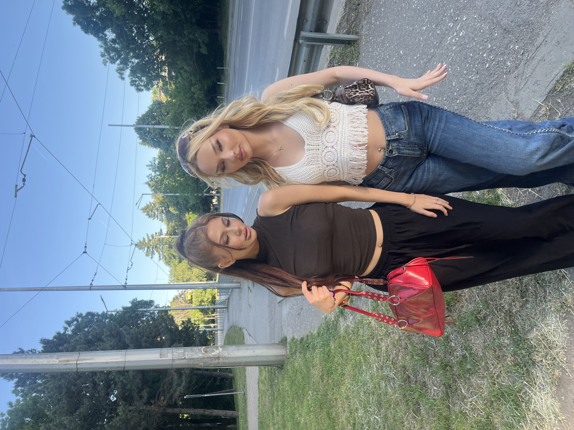 Two young women are posing on the edge of a sidewalk and grassy area next to a road. The woman closer to the camera has long brown hair pulled back in a ponytail and is wearing a brown tank top and black pants. She is holding a red handbag with a short chain strap. The other woman has long blonde hair and is wearing a white crop top and blue jeans with embellishments down the side. She is also carrying a small brown patterned purse. They both appear to be young adults. The location is likely Sofia, Bulgaria, due to the presence of tram lines and overhead cables, which are characteristic of the city's public transportation infrastructure. The background includes trees, a sidewalk, a road, and tram infrastructure. The sky is clear and blue, suggesting it is daytime.