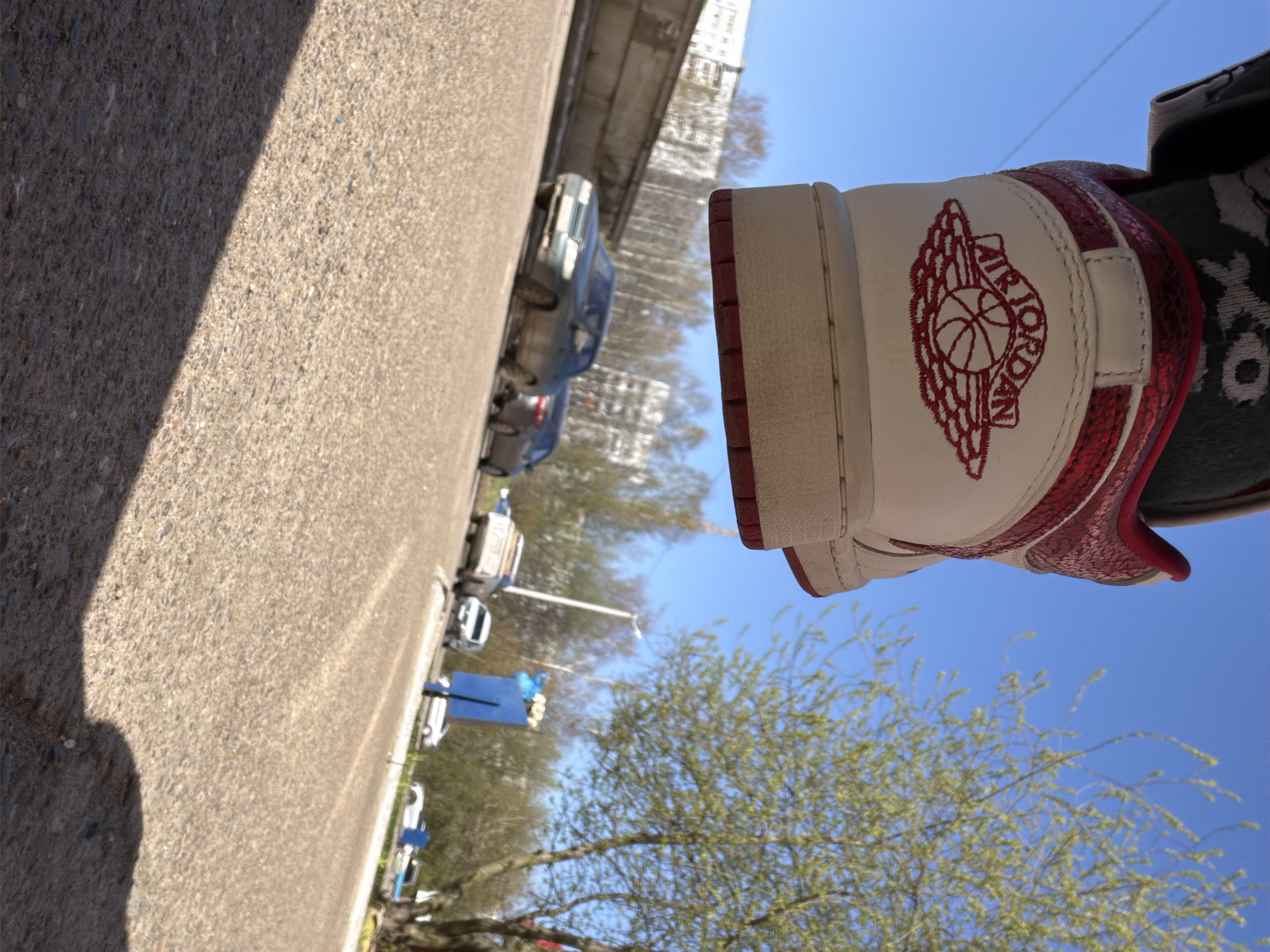 The image depicts a red and white Air Jordan sneaker in the foreground, positioned against a backdrop of a clear blue sky and a tree with budding green leaves. The sneaker's heel, featuring the Air Jordan logo in red embroidery, is prominently displayed.
In the background, a street scene unfolds, showing parked cars along the side of the road and buildings lining the street. The location is Chaikovsky, Russia.