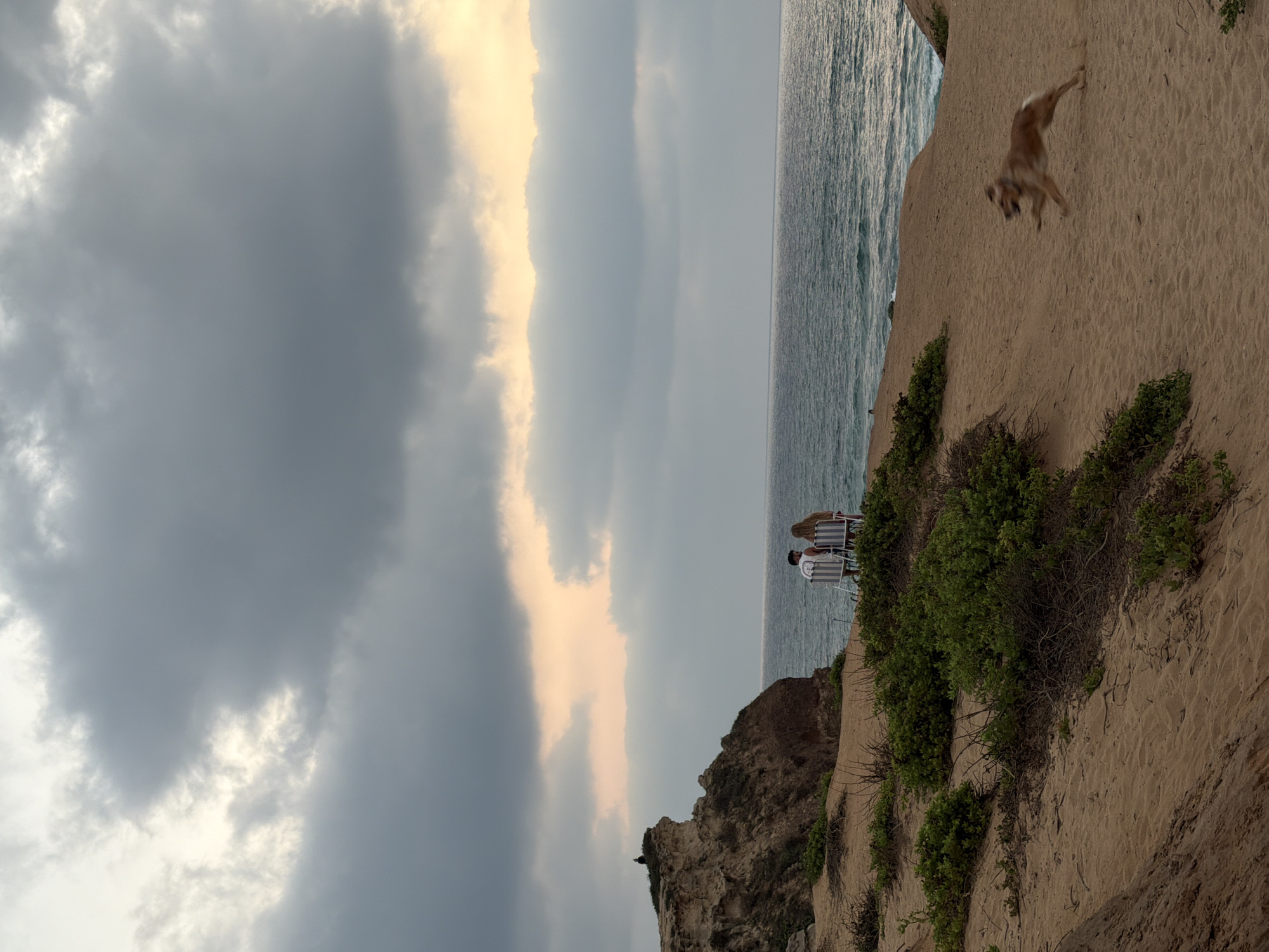 A couple sits in chairs on a sandy cliff overlooking the sea in Hadera, Israel. A dog runs across the sand in the foreground. The sky is filled with dramatic clouds, with a sliver of sunlight breaking through on the horizon. The sea is choppy, with waves crashing against the shore. The scene suggests a peaceful and natural setting.