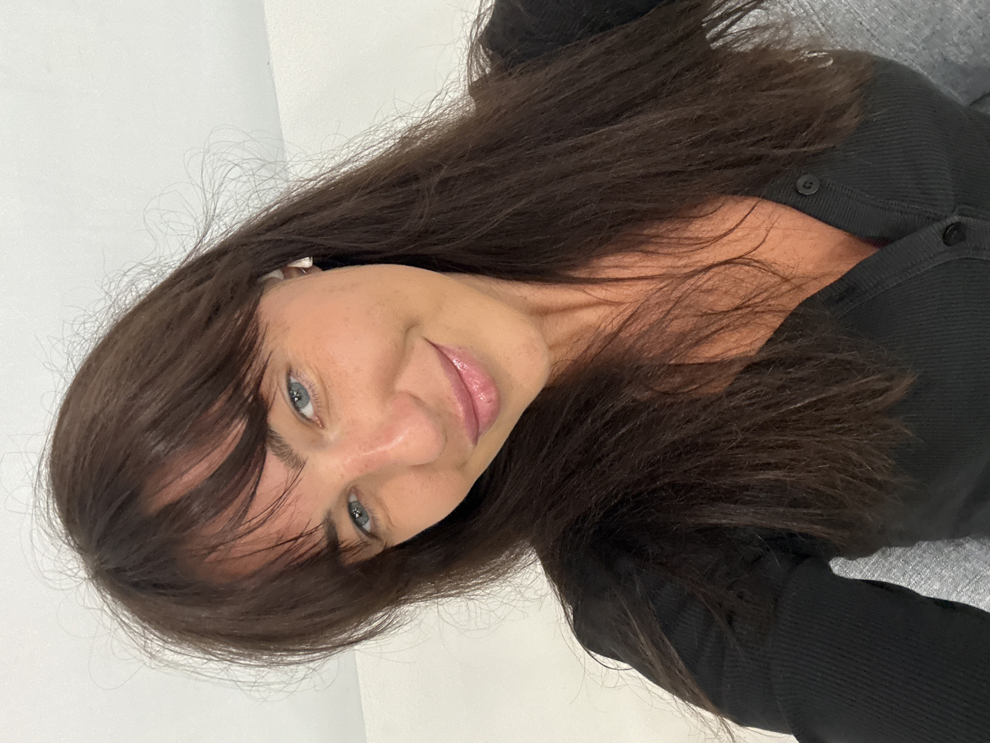 A woman with brown hair and blue eyes is photographed indoors. She is wearing a black top with buttons. The background appears to be a light-colored wall and a gray surface, possibly a couch or chair. The photograph is taken at an unusual angle.
