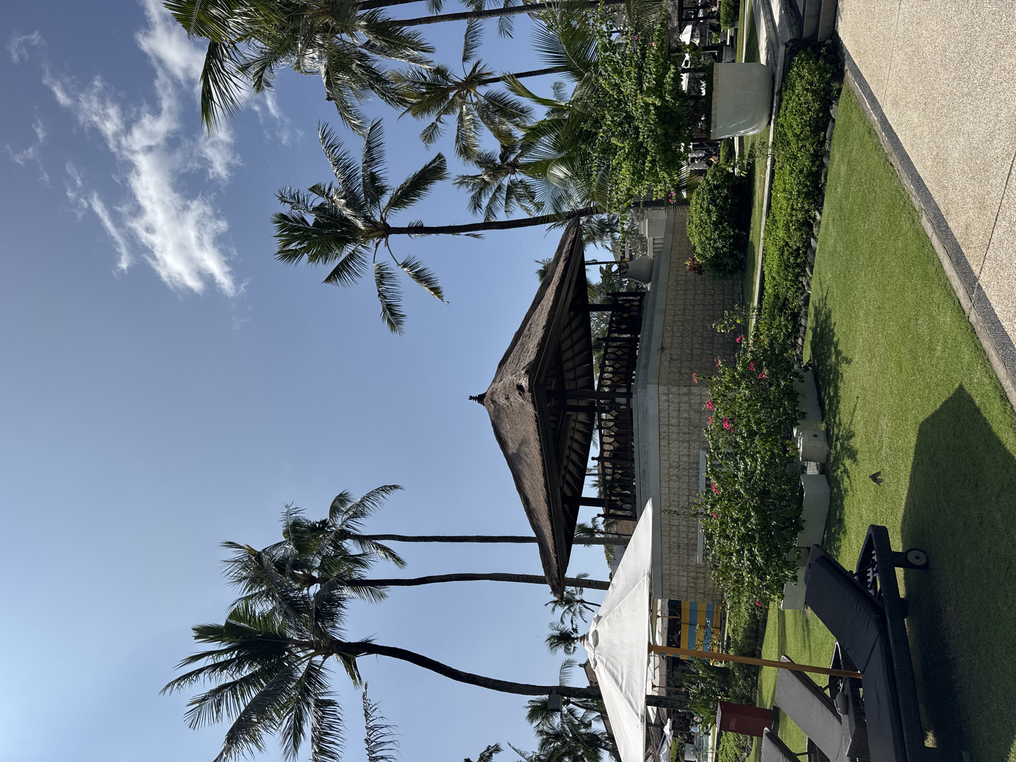 The image depicts a tropical resort scene in Kuta Selatan, Indonesia. The view is angled upwards, capturing tall palm trees against a blue sky with scattered clouds. A traditional thatched-roof pavilion stands amidst the palm trees. Adjacent to the pavilion, manicured greenery and flower beds border a grassy lawn. Sun loungers are positioned on the grass, partly shaded by a white umbrella. A paved walkway runs along the edge of the lawn. The overall impression is a serene and luxurious tropical setting.