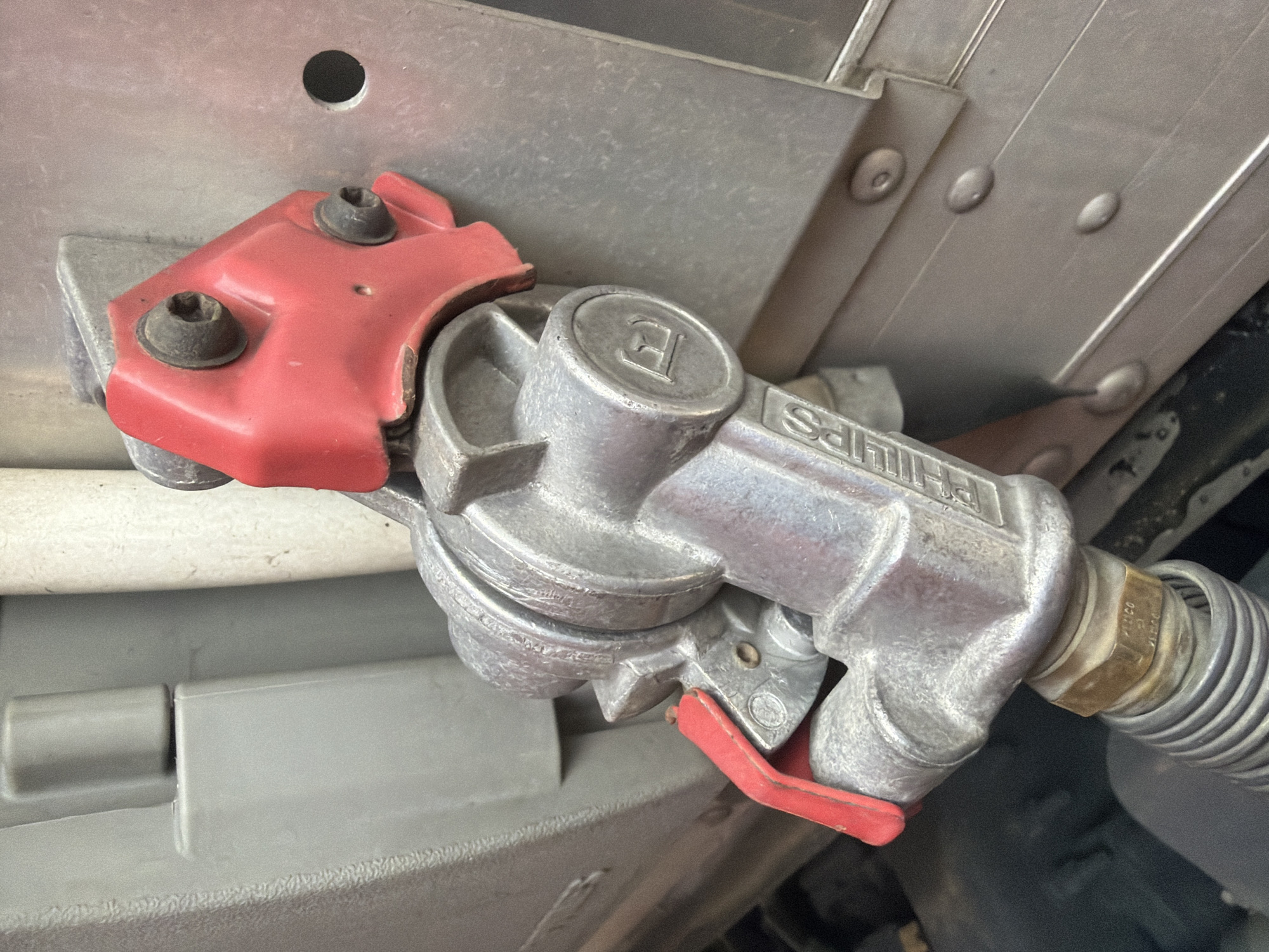 The image is a close-up, low-angle shot of a refueling nozzle with a red lever, attached to a metal conduit. The nozzle is silver and has the word "PHILIPS" embossed on it, along with a circular emblem containing the letter "E." The nozzle is connected to a corrugated hose at the bottom. Behind the nozzle, a section of a gray, textured surface is visible, possibly part of a vehicle. To the right of the nozzle, a silver metal panel with horizontal ridges and several circular rivets is present, consistent with the exterior of a truck or trailer. A small, circular dark hole is visible in this panel. The overall scene appears to be outdoors, possibly in an industrial or service area, but no specific location in Murfreesboro, United States of America, can be determined from the image alone. There are no people or discernible actions occurring in the frame.