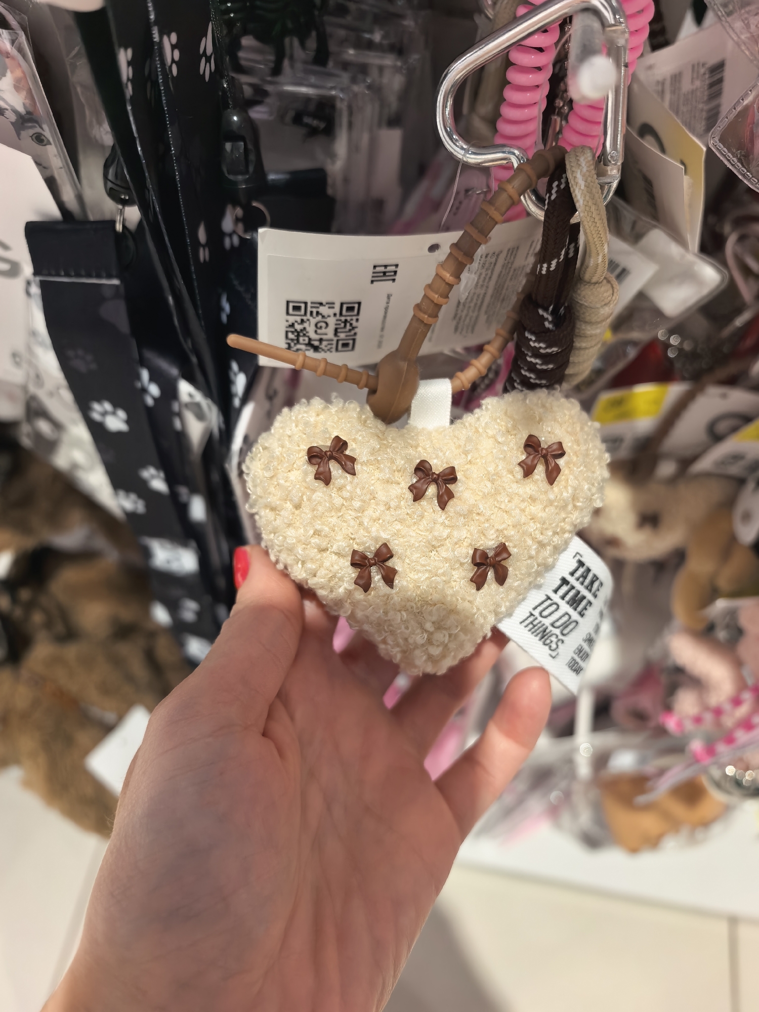A person's hand with bright pink nail polish is holding a fluffy, cream-colored heart-shaped keychain with small brown bows attached. The keychain is hanging from a metal clasp and a braided cord. A tag attached to the keychain reads "TAKE TIME TO DO THINGS, ENJOY TODAY". The background shows a display of various retail items, including lanyards with paw prints and other accessories. This image was taken in Sterlitamak, Russia.
