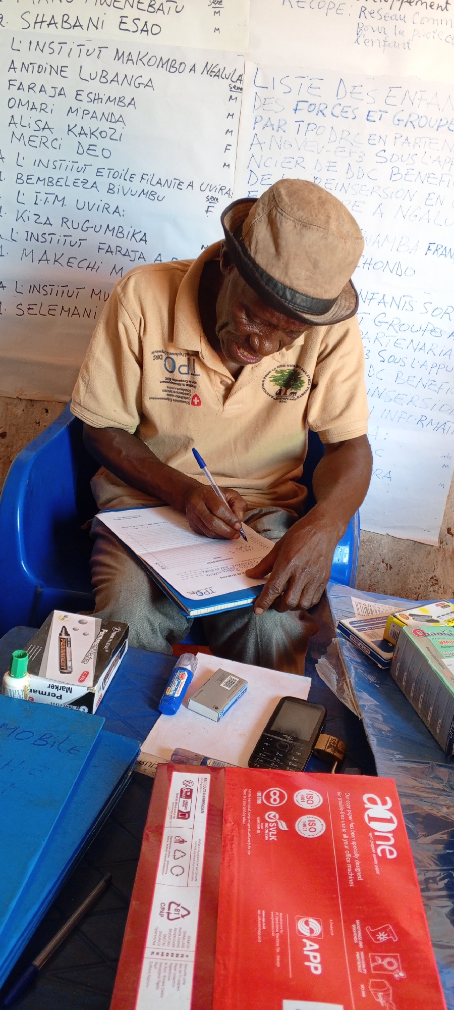 In Ngalula, Democratic Republic of the Congo, an elderly man with dark skin and wearing a tan shirt and hat sits on a blue plastic chair and writes in a notebook with a blue pen. The man is signing a paper. A table in front of him contains markers, glue, a cell phone, and paper. Two posters in the background list names and mention organizations working with children and reintegration.
