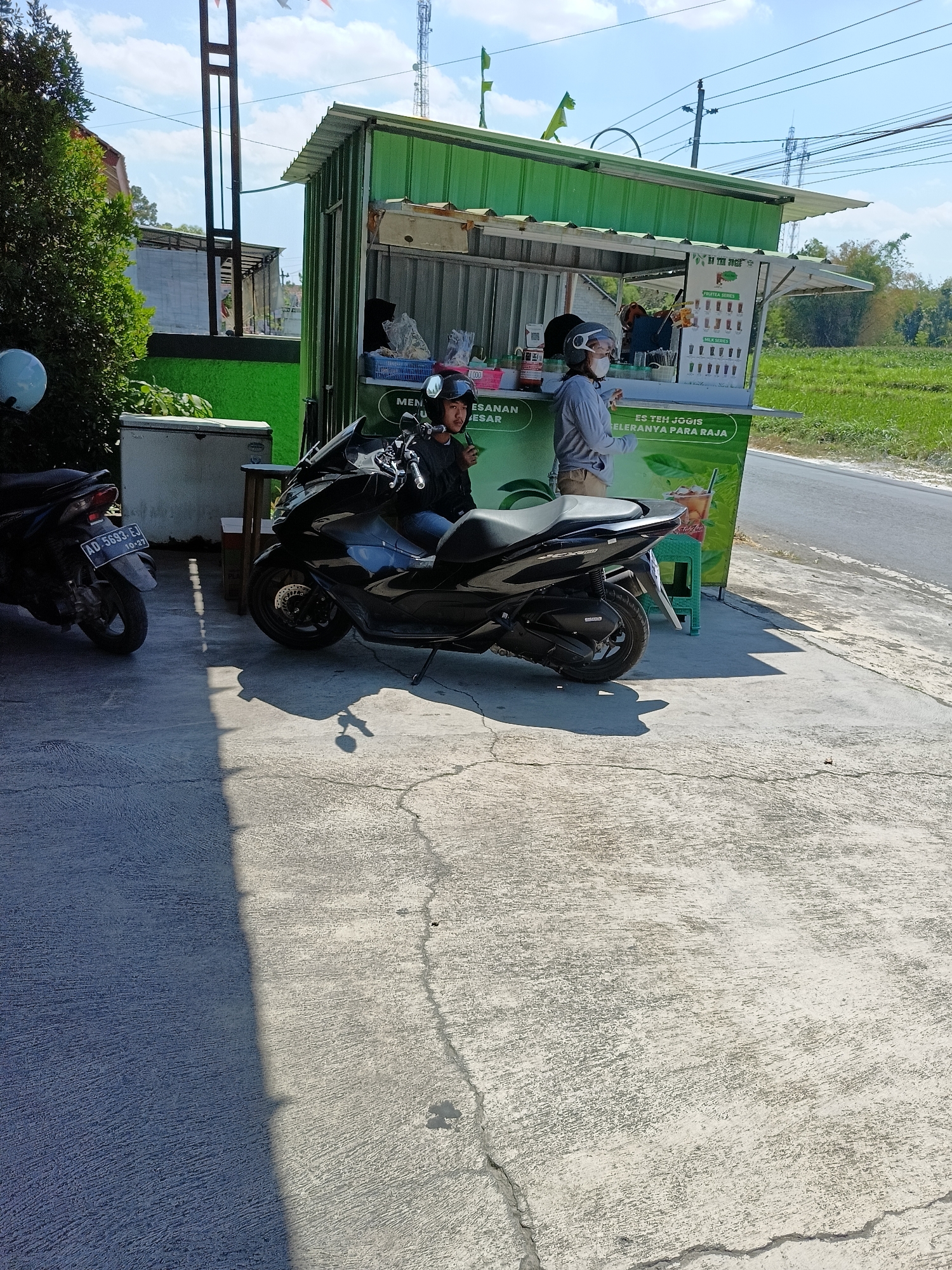 In Manisrenggo, Indonesia, a street-side beverage stand operates under a green corrugated metal awning. Two individuals are present: one wearing a black helmet and a gray long-sleeved shirt, sitting on a black motorcycle. The other, also wearing a helmet and a gray long-sleeved shirt, stands behind the counter. A menu board displaying "FRUIT TEA SERIES" and "MILK SERIES" is visible, along with various drinks. A white refrigerator sits to the left of the stand. Two black motorcycles are parked on a concrete surface, with one parked in front of the stand and another further to the left. The environment is illuminated by sunlight, casting shadows on the ground. A grassy field and trees are visible in the background, with power lines and a cell tower in the distance.