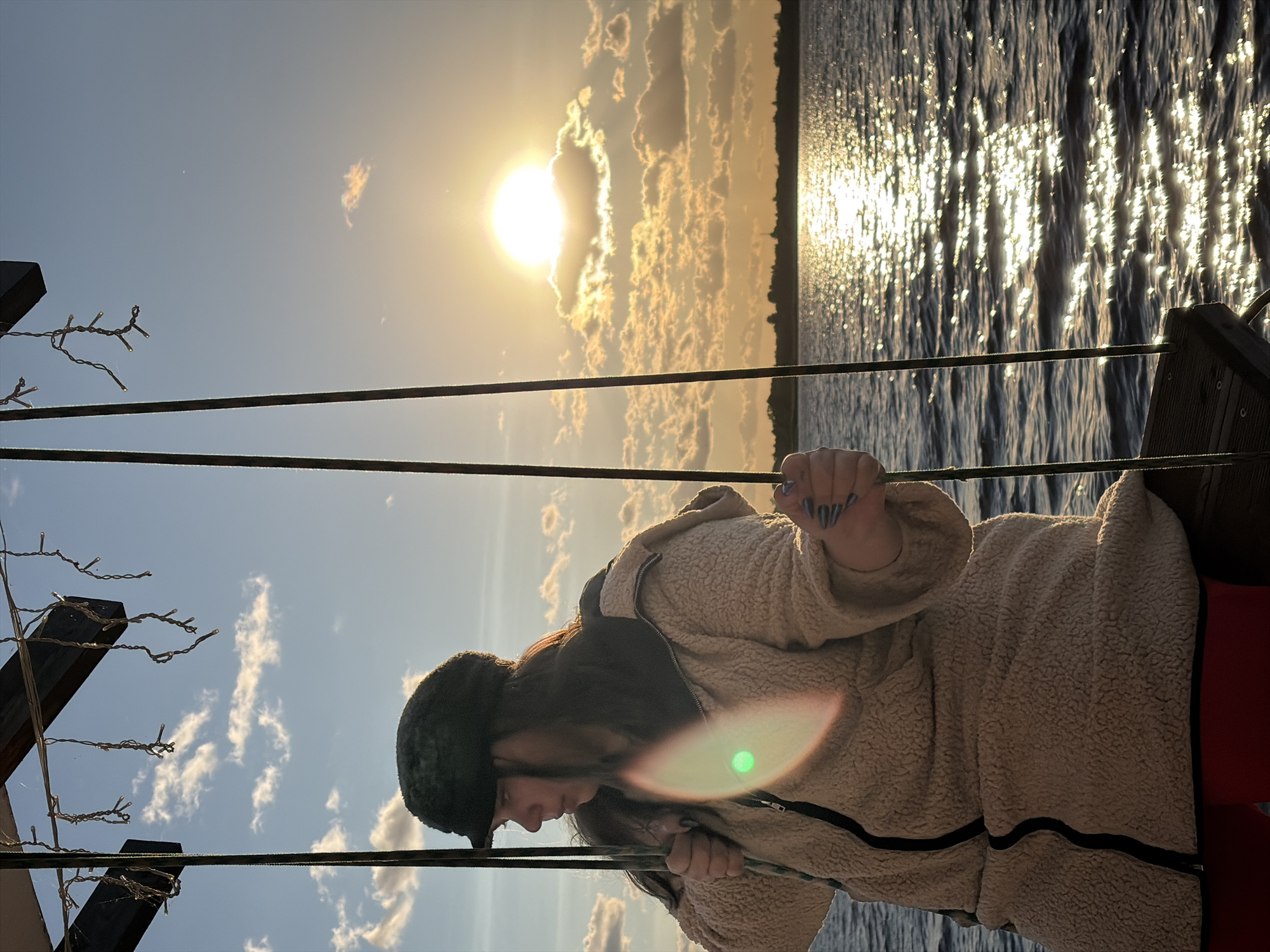 A person sits on a wooden swing suspended by ropes, facing away from the camera and looking towards the right. The person is wearing a fuzzy, light-colored jacket with a dark zipper and a dark cap. The background features a body of water with sunlight reflecting off its surface, a distant tree line, and a cloudy sky with the sun setting. The scene appears to be outdoors, possibly a waterfront area in Vidogoshchi, Russia, given the context.