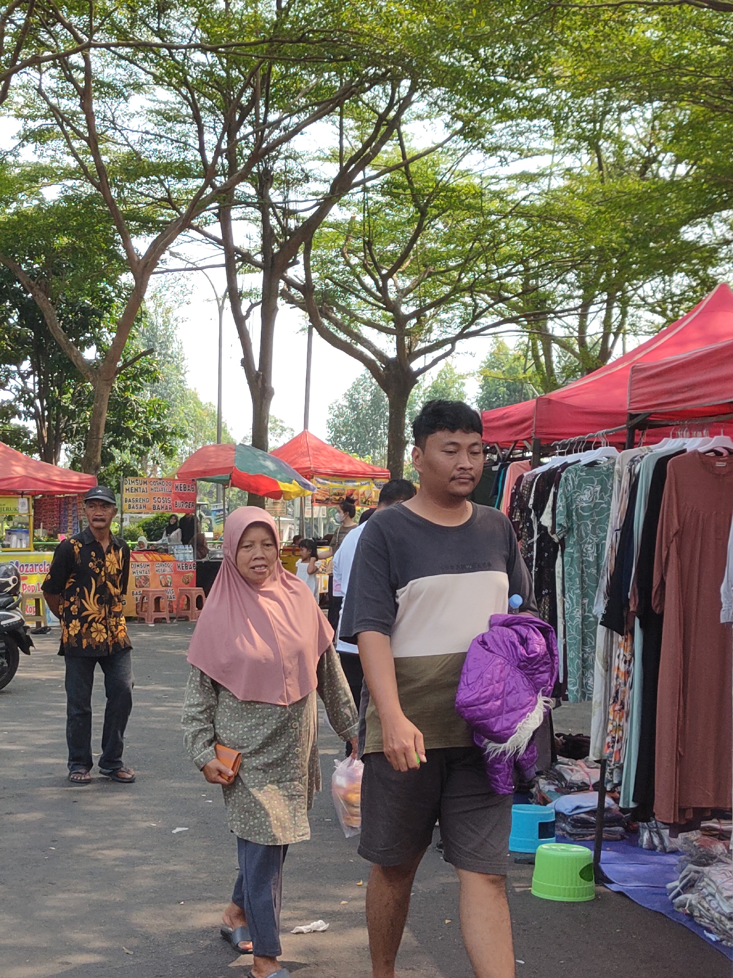 A street market scene is captured in Ngasem, Indonesia, with a focus on individuals browsing or walking through. In the foreground, a man in a black and gold patterned shirt and dark pants, wearing a black cap and flip-flops, stands looking to his left. Beside him, a woman wearing a pink hijab and a long patterned dress with a green and white floral design, walks forward holding an orange wallet in her right hand and a plastic bag in her left. To their right, a man in a gray, white, and brown t-shirt and dark shorts carries a folded purple jacket over his left arm. Behind them, various market stalls are visible, including one with red awnings displaying racks of clothes for sale. Other individuals and market activity are present in the background, under the shade of large trees. The ground is paved, and sunlight filters through the leaves.