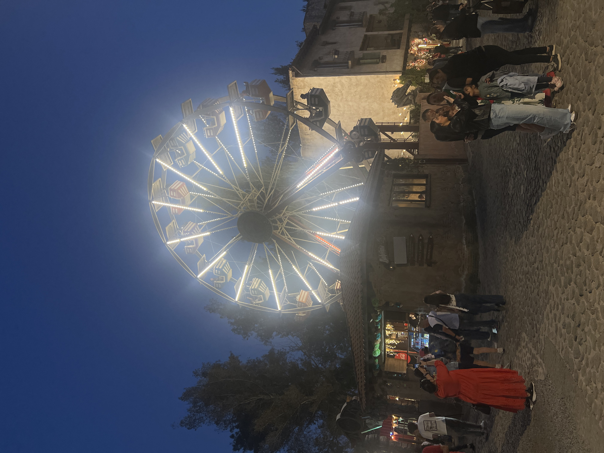 A Ferris wheel illuminated with bright lights is the primary focus of the image, situated against a darkening blue sky. The surrounding area appears to be a cobblestone street in the city of Domingo Arenas, Mexico, with buildings on one side. There are people standing and walking on the street. Some individuals are taking pictures of the Ferris wheel. The environment suggests an evening or nighttime setting, potentially a tourist spot.