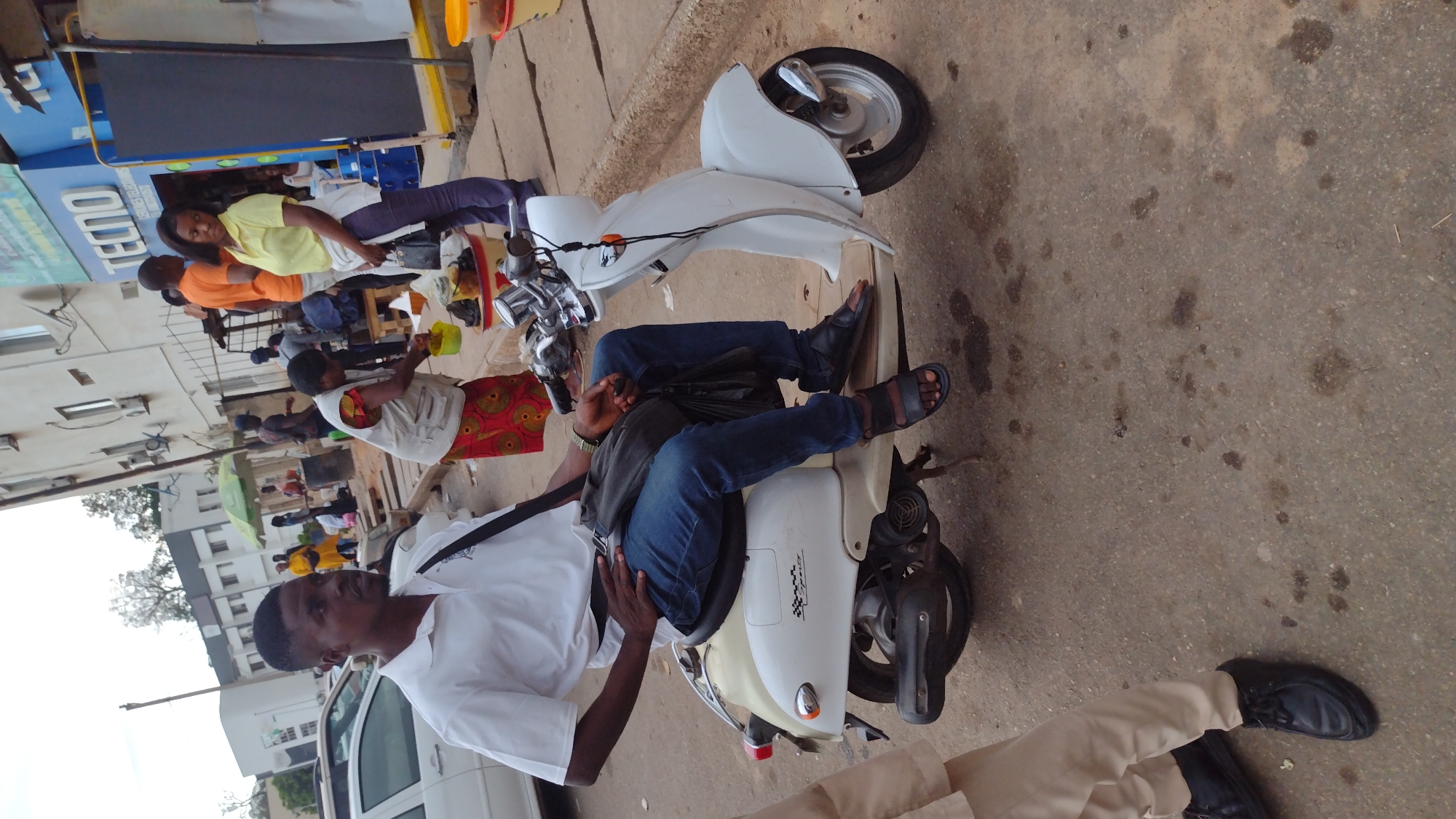 The image captures a street scene in Jos, Nigeria. A man in a white shirt and black trousers is standing near a white moped. Another individual, wearing jeans, a gray top, and sandals, is reclining on the moped. Behind the man in white, there is a group of people near a table, one wearing an orange shirt, another a yellow shirt, and another an apron with a colorful patterned skirt. Buildings are visible in the background, including a shop with a blue sign with the name “TECNO” and a multi-story building further back. A parked white car is also visible on the left side of the image. The ground appears to be paved with concrete and has some discoloration.