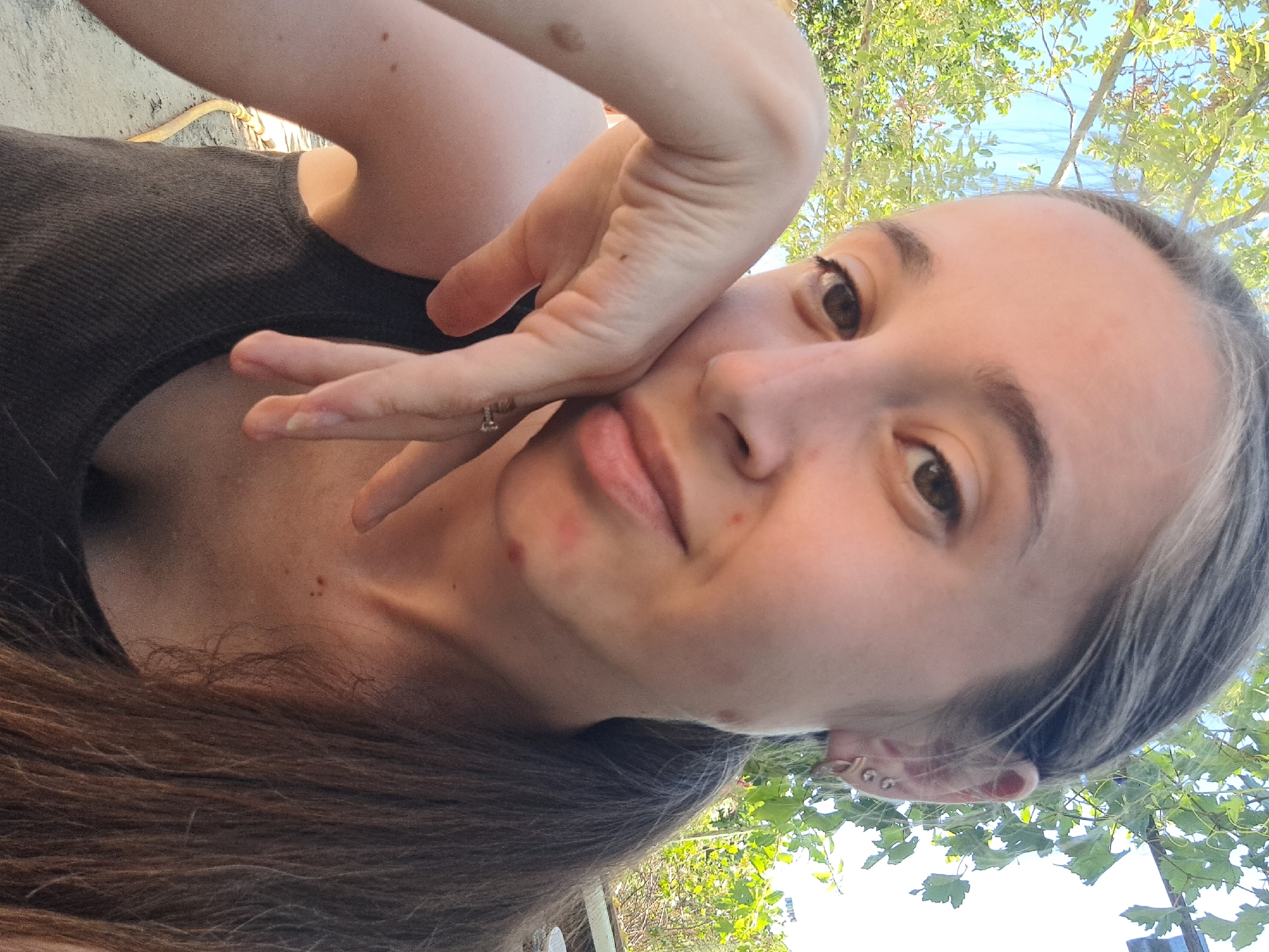 The image shows a close-up, selfie-style photograph of a young woman outdoors. She has long, brown hair, pulled back into a ponytail, and is wearing a dark tank top. Her left arm is propped up, with her hand resting on her cheek. She is wearing a ring on her right hand and earrings in her left ear. The background consists of green foliage and a bright sky, suggesting a daytime setting. There are no indications that the photograph was taken in Vypasne, Ukraine, nor are there any other elements in the image that suggest a specific urban or geographical location. The image focuses entirely on the woman and her immediate surroundings, with no identifiable landmarks or contextual clues about her location.