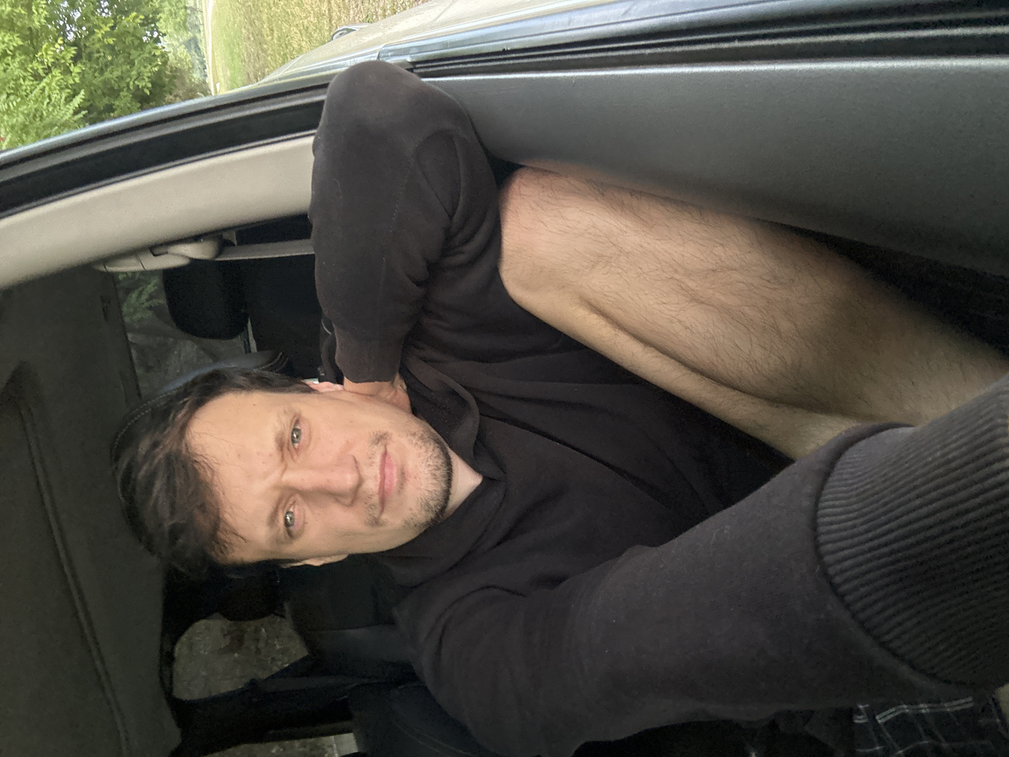 A man is sitting in the passenger seat of a vehicle, looking towards the camera. He has dark hair and a light complexion, with a day's growth of stubble on his chin and jawline. He is wearing a black hooded sweatshirt and his left arm is resting on the open window frame. His right leg is visible, and it appears to be bare. The interior of the vehicle is dimly lit, with shadows cast by the overhead compartment and rear seats. The exterior of the vehicle shows a grassy area with trees in the background. The scene appears to be outdoors, possibly in a rural or suburban setting. There is no explicit information available to specify the exact location within the city of Makiv, Ukraine.