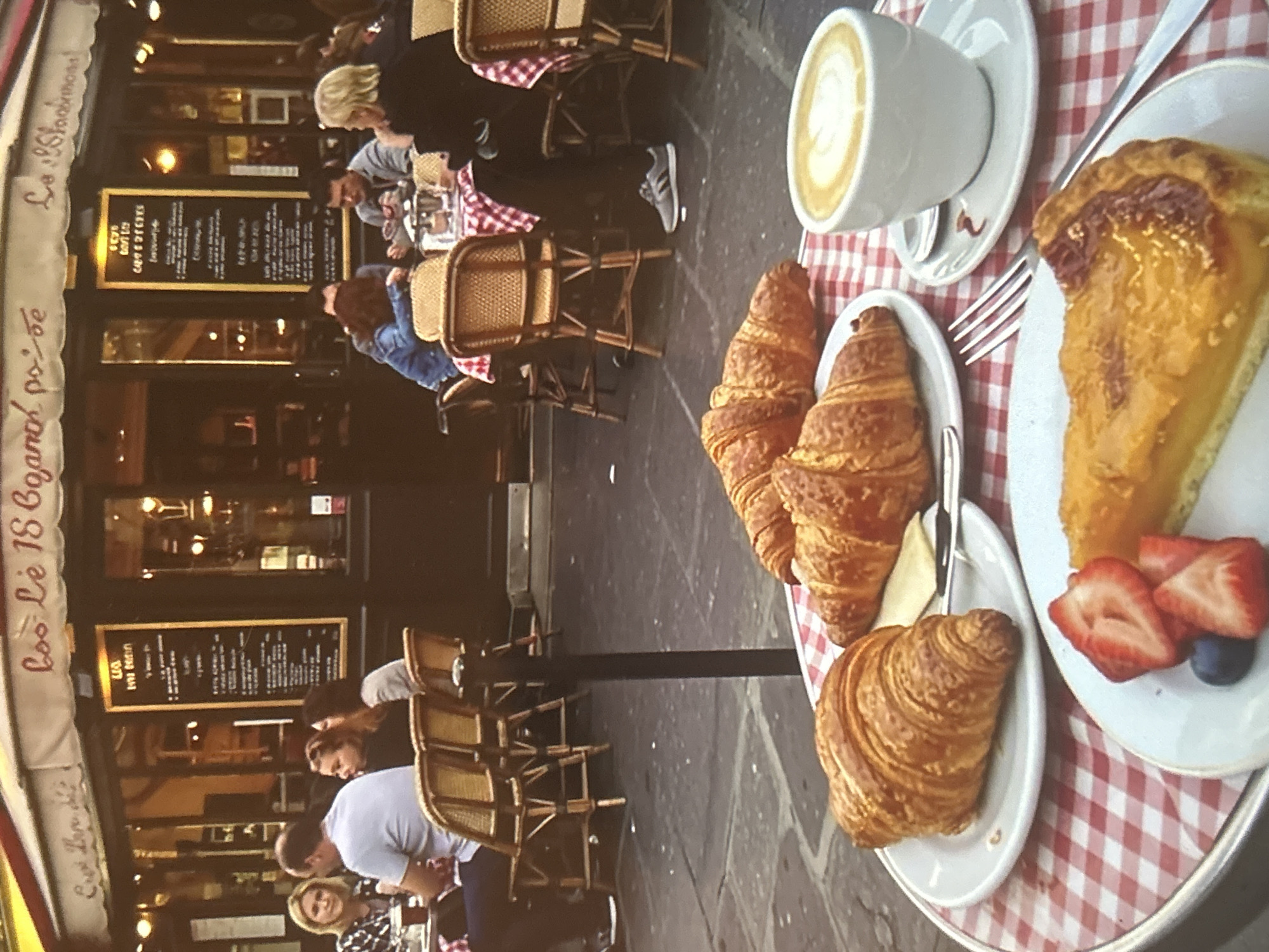 The image captures a close-up view of a table at an outdoor cafe with a checkered tablecloth. On the table, there are two croissants on a white plate, a slice of fruit tart garnished with strawberries and a blueberry on another white plate, and a white cup of coffee with latte art, placed on a saucer with a spoon. In the background, several people are seated at tables outside a cafe with awnings and menus displayed on the windows. The cafe has a sign with French text. The setting appears to be a European city.