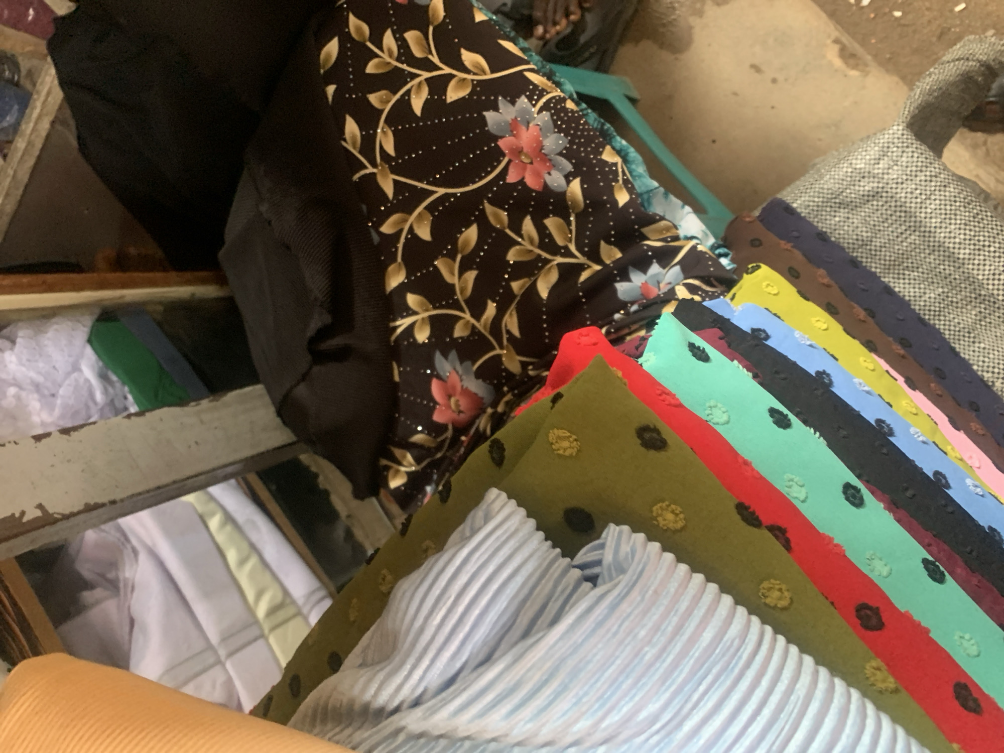 This is a top-down, eye-level shot of a collection of fabrics, likely in a market or shop setting in Jalingo, Nigeria. The dominant subjects are various textiles, arranged in stacks and folds. In the foreground, a roll of orange, ribbed fabric is visible, along with a section of light blue, pleated fabric. Behind these, a series of colorful, textured fabrics are laid out diagonally, including olive green with yellow polka dots, red, teal with black polka dots, light blue, yellow, and dark brown. Further back, a dark fabric with a floral pattern featuring pink and blue flowers and gold accents is prominent. To the far left, a glimpse of white and pale yellow folded fabrics can be seen within a rustic, wooden structure that resembles a shelf or display case. A piece of gray and black patterned fabric is visible on the right side. The scene appears to be indoors or under cover, given the lighting and the presence of a concrete floor with some debris. There are no people directly visible, but a hint of a person's leg in dark clothing is visible towards the top right, suggesting a person might be nearby, perhaps a vendor or shopper. The overall impression is one of vibrant colors and textures, hinting at a place where textiles are sold or displayed.