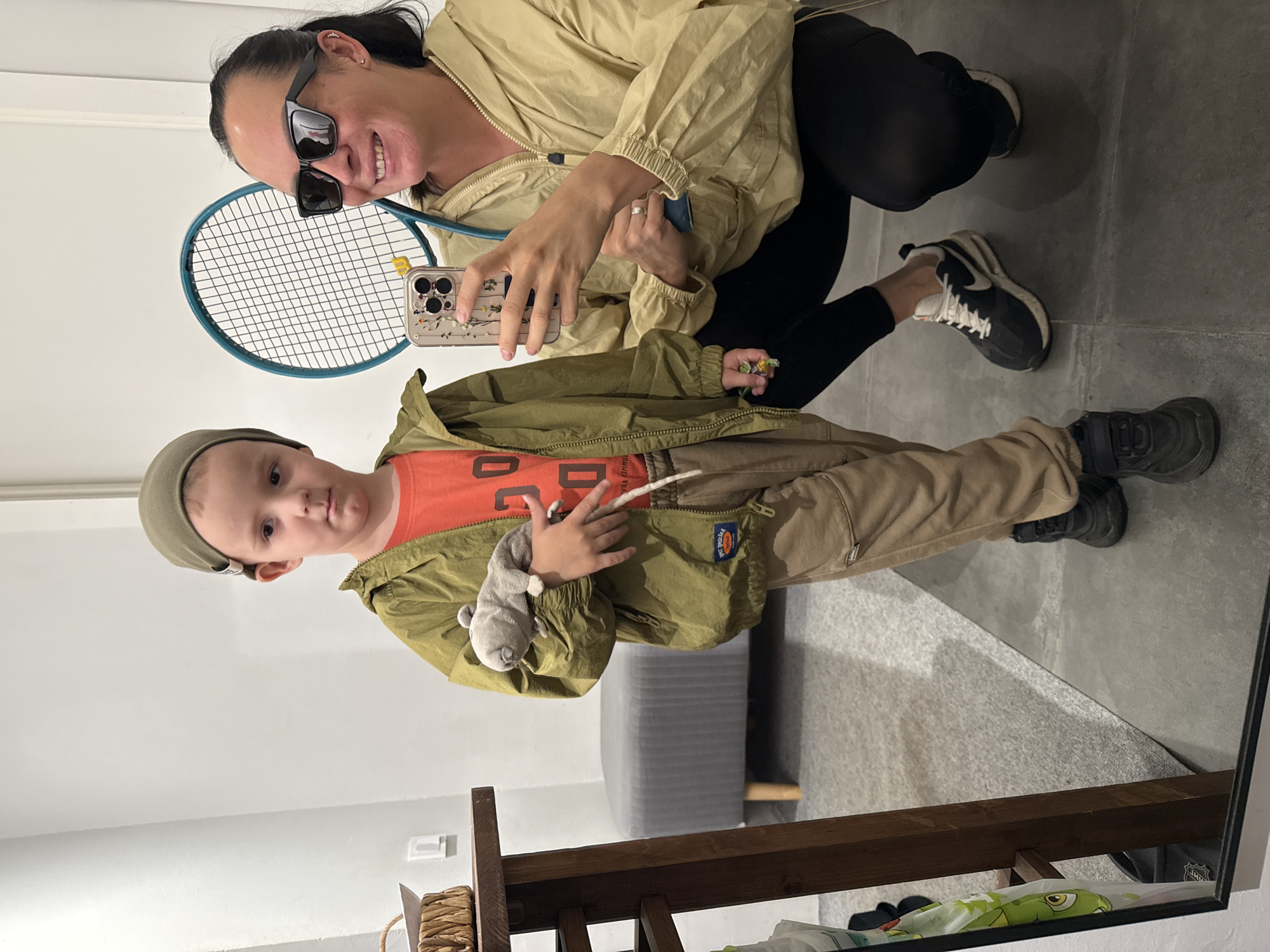 The image shows a young boy and a woman posing for a selfie. The boy is wearing a green jacket, an orange t-shirt, and tan pants, and he is holding a stuffed mouse. The woman is wearing a light yellow jacket and black pants. She is wearing sunglasses and holding a phone to take the picture. A tennis racket is visible in the background. The setting appears to be an indoor space with a mirror, suggesting a fitting room or a similar retail environment. The floor is tiled, and there is a stool and a wooden table with a basket in the background. The overall lighting is bright. No specific landmarks or indications of Chaikovsky, Russia are present in the image.