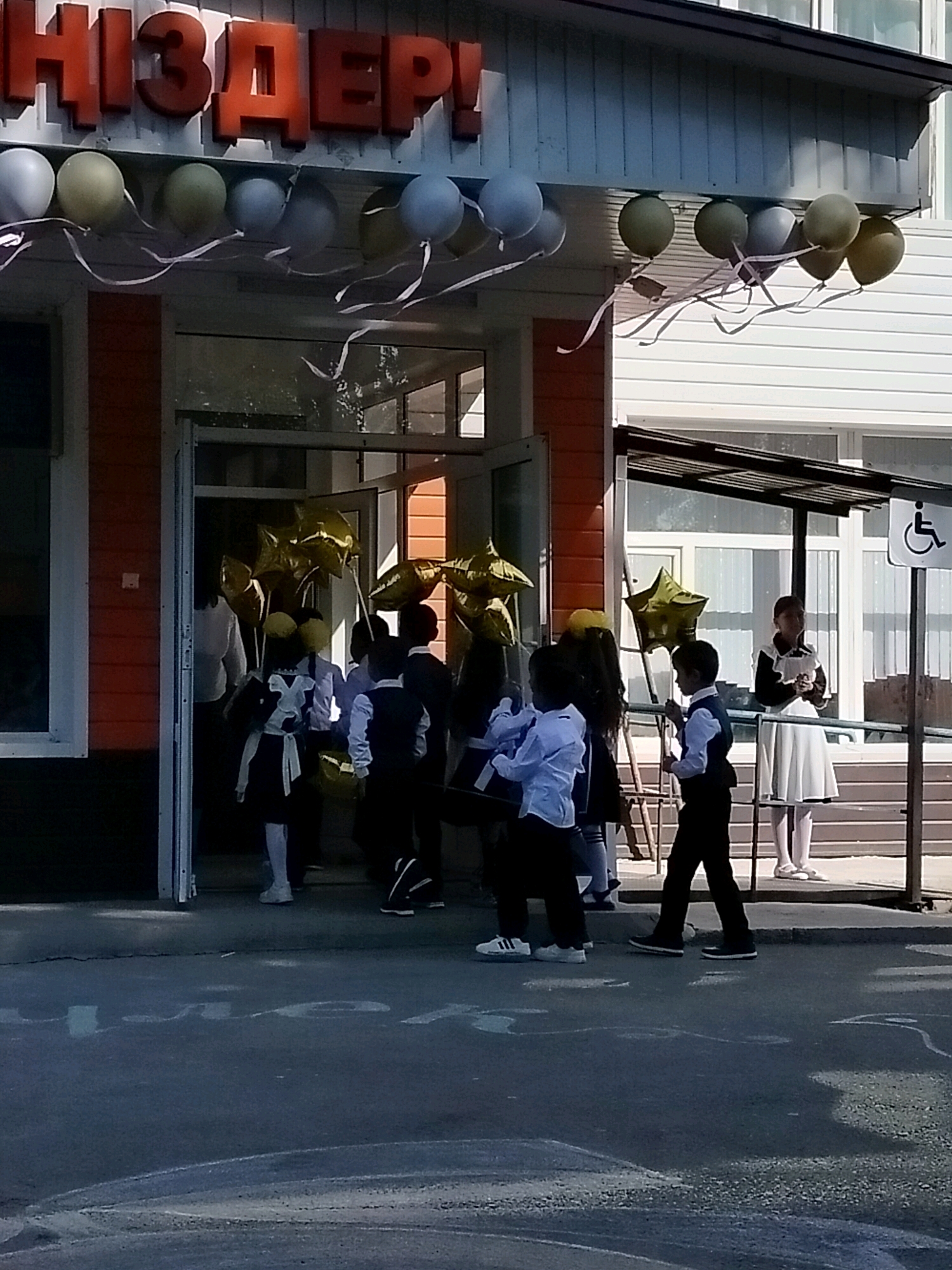 The image depicts the entrance of a building with red and white siding, adorned with balloons. The building's facade features large, red, three-dimensional letters spelling out "НІЗДЕР!". A row of balloons in various colors, including light blue, olive green, and yellow, are attached to the building with white ribbons. Some of these balloons are star-shaped.
Several children in school uniforms are seen entering and exiting the building through an open doorway. The uniforms appear to consist of dark skirts or trousers and light-colored shirts, with some boys wearing dark vests. One child in the foreground is wearing a dark skirt with a white ribbon tie. Another child, a boy, is wearing a dark vest over a light shirt and dark trousers.
In the background, a woman in a white dress and a dark top stands near a window, holding a piece of paper. A sign with a wheelchair symbol is visible on a post to the right of the entrance. The ground in front of the building is paved with asphalt, and chalk markings are visible on the surface. The overall scene suggests a school or an educational institution, possibly decorated for an event such as the first day of school or a celebration. The setting is outdoors, with daylight visible, indicating a daytime scene. The architectural style of the building and the presence of the Cyrillic alphabet in the signage suggest a location within a region where Cyrillic is used. The city of Көлкент, Kazakhstan, is a plausible location given this context.