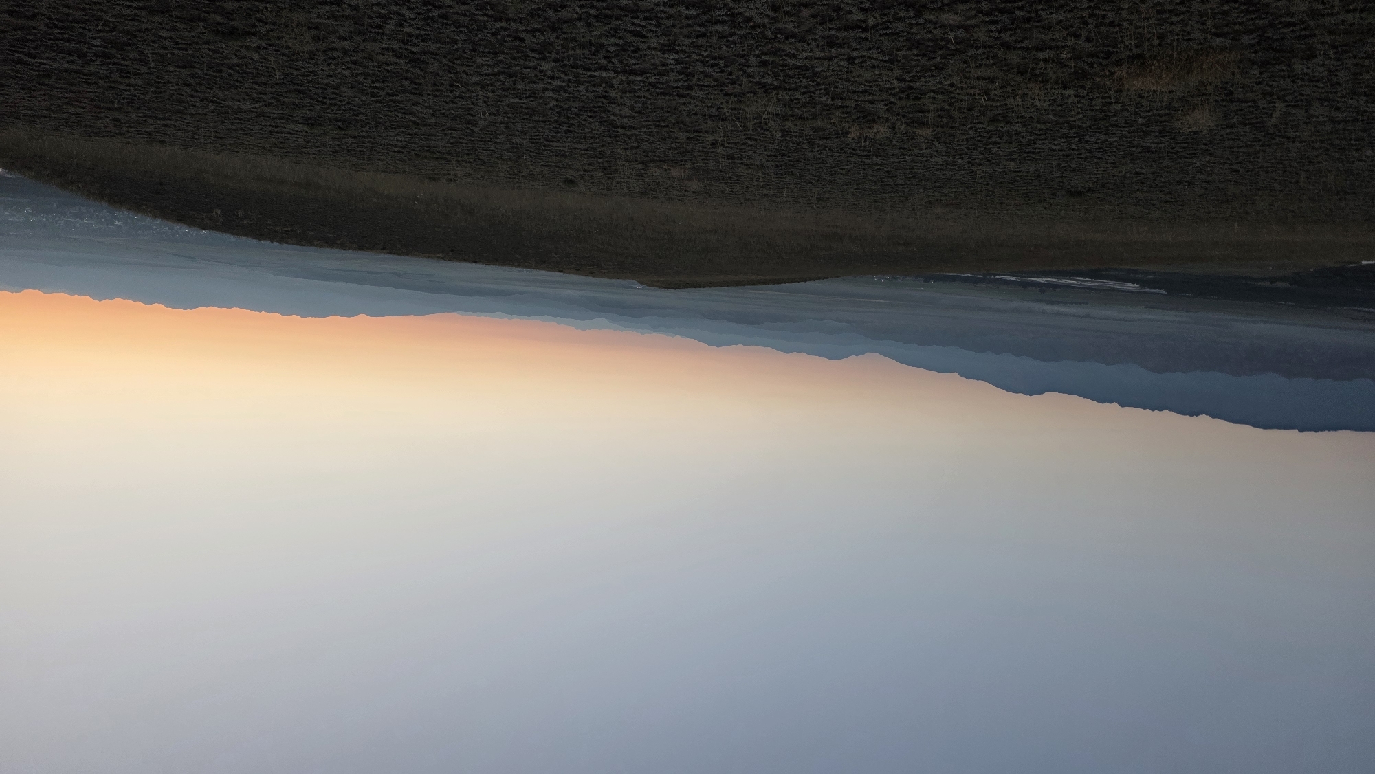 The image captures a wide, panoramic view of a mountainous landscape at dawn or dusk. The sky transitions from a pale blue at the top to a warm gradient of orange and pink near the horizon. A range of mountains, appearing layered and distant, stretches across the middle ground, their silhouettes softened by the atmospheric haze. In the foreground, a gently sloping hill is covered in dry, sparse vegetation, casting a dark shadow. The terrain appears to be a rural or natural setting, possibly on the outskirts of a settlement. The location is identified as Ак Жар, Kyrgyzstan. There are no people or animals visible in the image. The overall impression is one of serene natural beauty and vast open space.