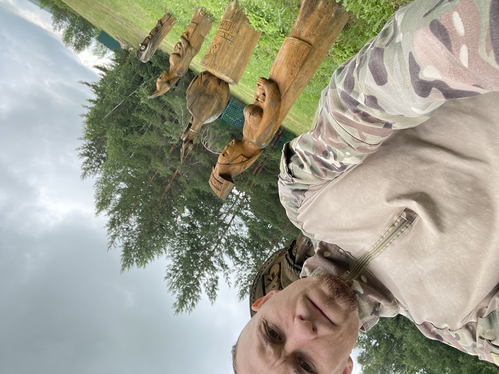 A man takes a selfie outdoors, holding the camera low to capture himself and several wooden sculptures. He is wearing a camouflage-patterned long-sleeved shirt. The sculptures, carved from logs, are in a line on the grass, depicting figures with human-like features. Behind the sculptures, a dense forest of evergreen trees is visible under a cloudy sky. The ground is covered in green grass and some smaller plants. The image appears to be taken in a rural or park-like setting in the city of Nikolayevka, Russia.