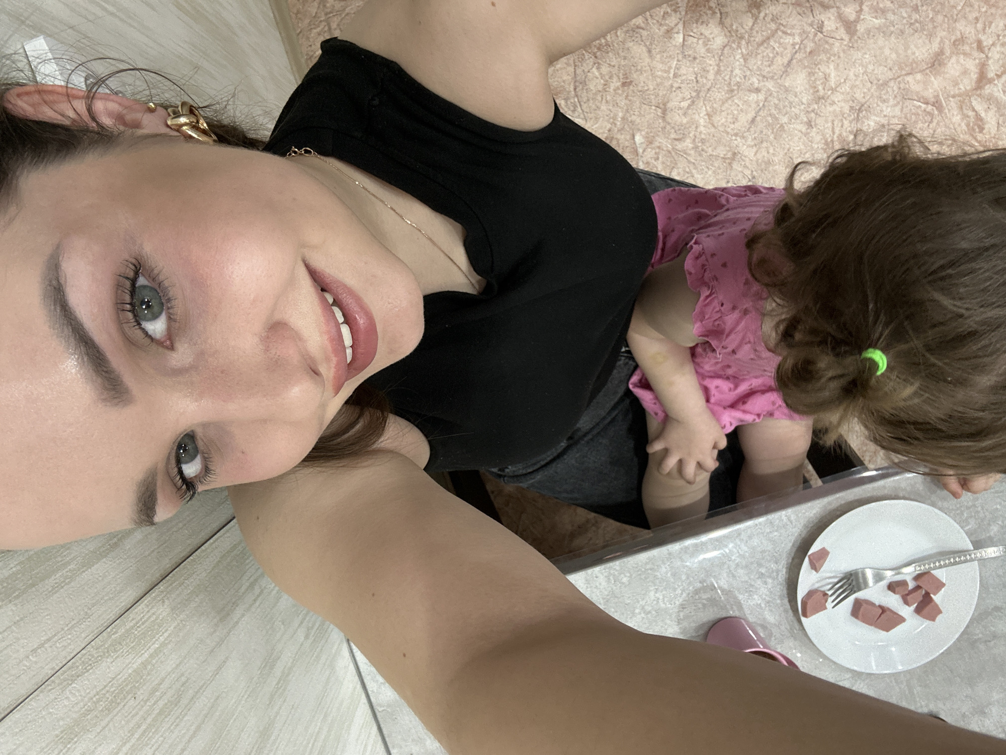 A woman and a child are in a room. The woman is fair-skinned with dark hair and is wearing a black top and gold earrings. The child is wearing a pink dress. On a plate in front of the child are several pieces of sausage, cut up, along with a fork. A small pink shoe is also visible next to the plate. The floor is made of light-colored wood or laminate. A textured beige fabric is visible in the background. The location is Chaikovsky, Russia.