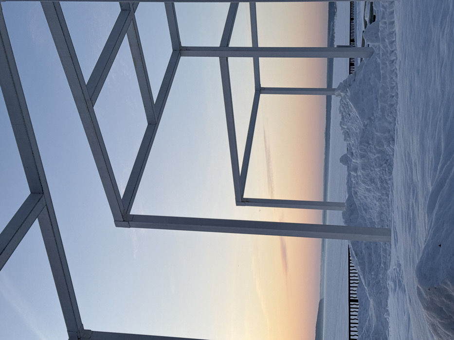 The scene captures a winter landscape on an embankment overlooking the Kama Reservoir in Chaikovsky, Russia. The foreground is covered in a substantial layer of pristine snow, featuring several uneven piles and drifts. Three distinct white, open-frame cubic or rectangular structures, composed of square-section metal beams, are prominently featured in the midground; one is partially visible on the left, another centrally positioned, and a third further back on the right. A dark, low-level railing or fence runs horizontally behind the snow piles, marking the edge of the elevated ground. Beyond this, a vast, flat expanse of frozen water, the Kama Reservoir, stretches to the distant horizon. A faint, snow-covered landmass is discernible on the far left. The sky above transitions from a pale blue directly overhead to a soft pink and orange gradient at the horizon line, indicative of dawn or dusk. No individuals, activities, or interactions are visible within the frame.