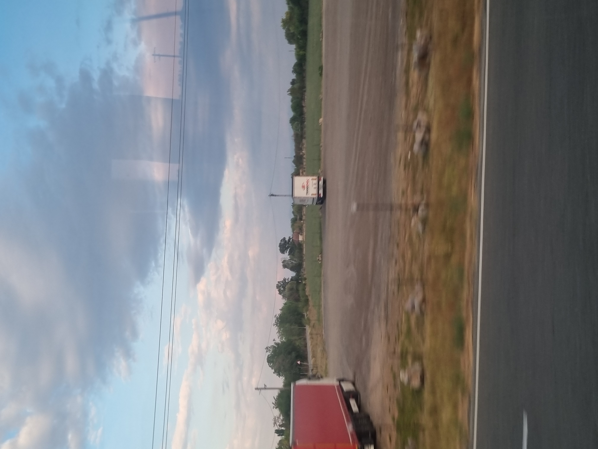 The image depicts an outdoor scene, likely a roadside or parking area, with two trucks visible. The truck on the left is predominantly red and white, and its rear is facing the camera. The truck on the right is white with some text and graphics on its side, and it is also facing away from the camera. Both trucks appear to be stationary. The ground is composed of gravel and dirt. In the background, there is a line of trees and vegetation. The sky is partly cloudy with shades of blue, gray, and hints of pink. There are power lines running horizontally across the upper portion of the image. The foreground shows a section of an asphalt road with a white stripe. The scene suggests a rural or semi-rural location. It is difficult to ascertain the exact time of day, but the lighting indicates it may be late afternoon or early evening. The location is identified as Mădăras, Romania.