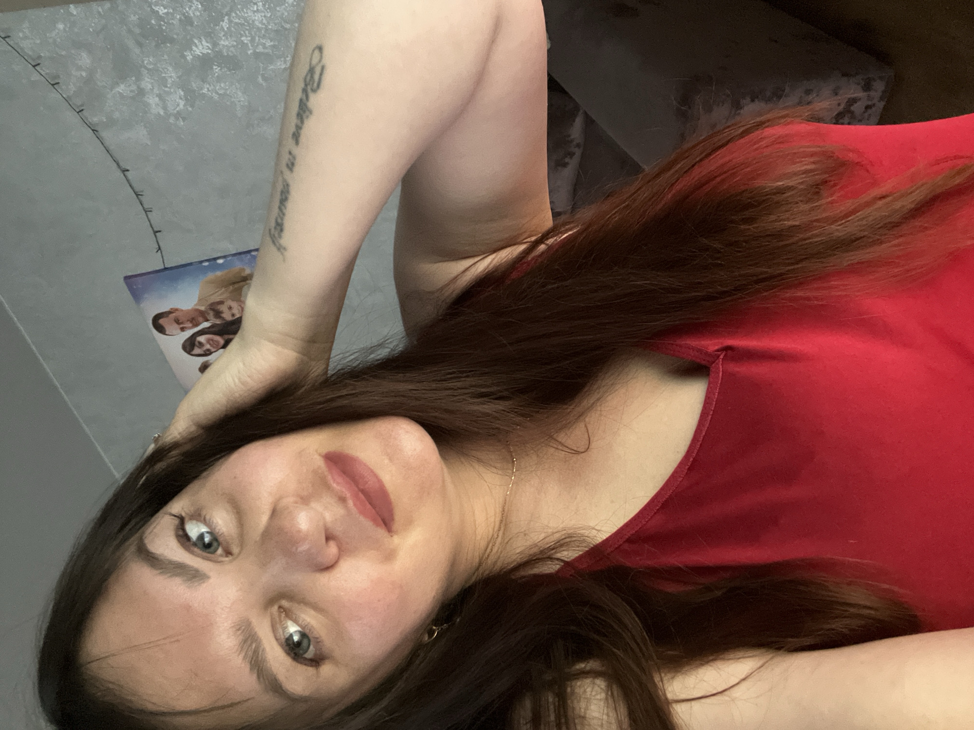 A young woman with long, dark brown hair and light blue eyes is taking a selfie in a room. She is wearing a red tank top and a delicate gold necklace. Her right arm is raised, and her hand is touching her hair. A tattoo is visible on her left forearm, which reads "Believe in yourself." In the background, a framed picture of a family and a string of small lights are attached to a textured grey wall. A dark grey couch is partially visible on the right side of the image. There is no indication of the city of Noviy, Russia within the image itself.