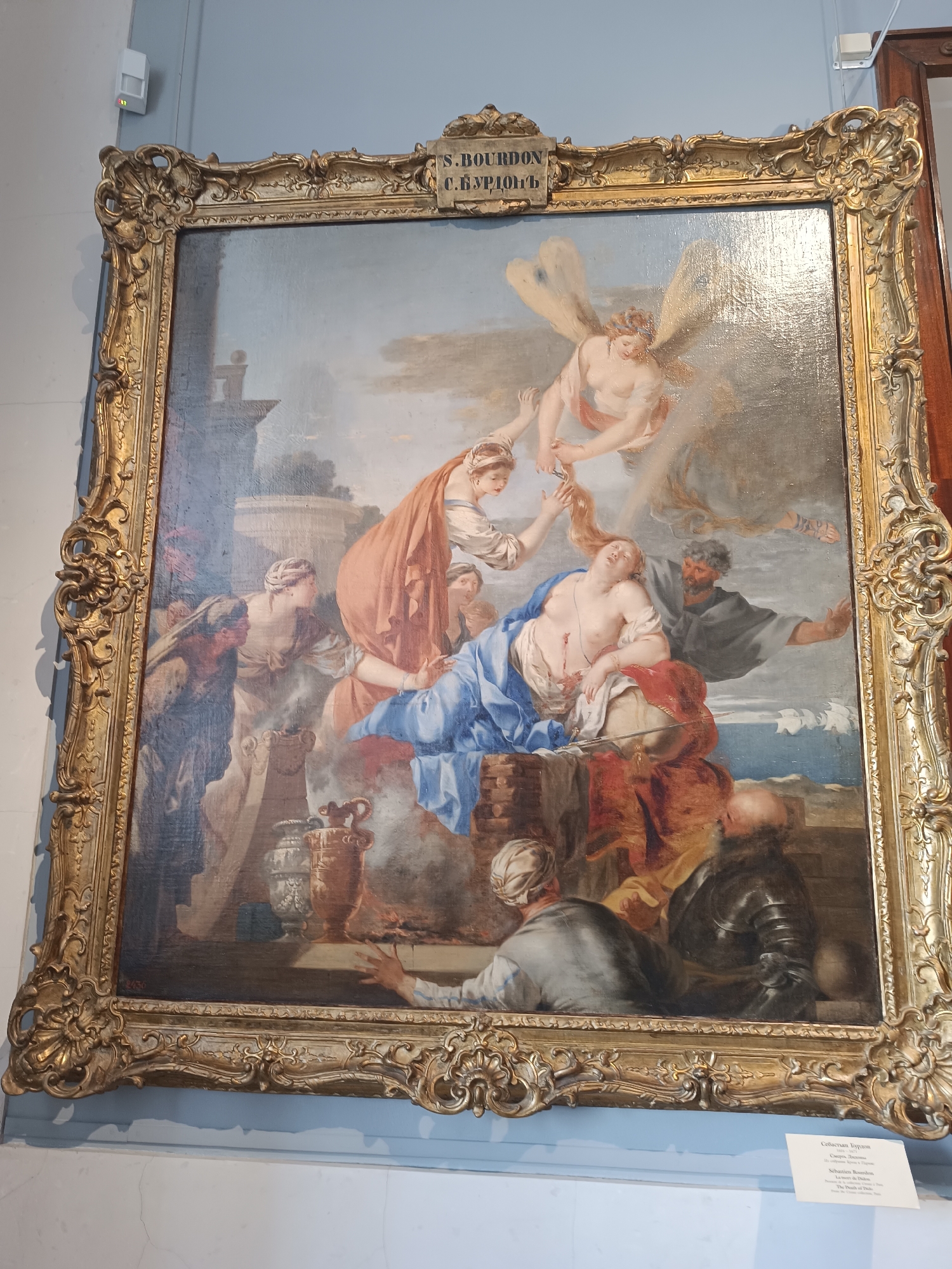 An oil painting in a gilded frame depicts a mythological or religious scene. A central female figure, appearing to be a young woman, reclines on a draped surface, her head tilted back, and her eyes closed. She is partially undressed, with her torso exposed, and there are visible wounds or marks on her chest. A winged cherubic figure descends from above, holding a pair of scissors as if to cut her hair. Another male figure, with a beard and wearing a dark robe, stands behind the reclining woman, reaching out a hand towards her. Several other figures are present in the scene, some gathered around the central figure, while others are in the background. One figure on the left, wearing a head covering, points towards the reclining woman. In the foreground, a man in armor looks on. The setting appears to be an outdoor or architectural space, with classical structures visible in the background. The overall impression is dramatic and emotional. The painting is displayed on a plain, light-colored wall. A small informational placard is visible at the bottom right corner of the image, partially obscured by the frame.