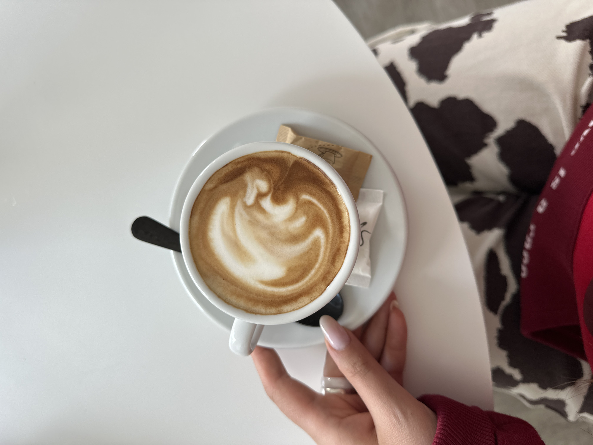 A top-down shot depicts a cup of latte art coffee resting on a white saucer, accompanied by a black spoon and two packets of sugar. The saucer sits on a white table. A hand with light pink nails and a silver ring is holding the cup. Part of a cow-patterned pillow and a red shirt sleeve are visible in the background. The location is the city of Bar, Montenegro.