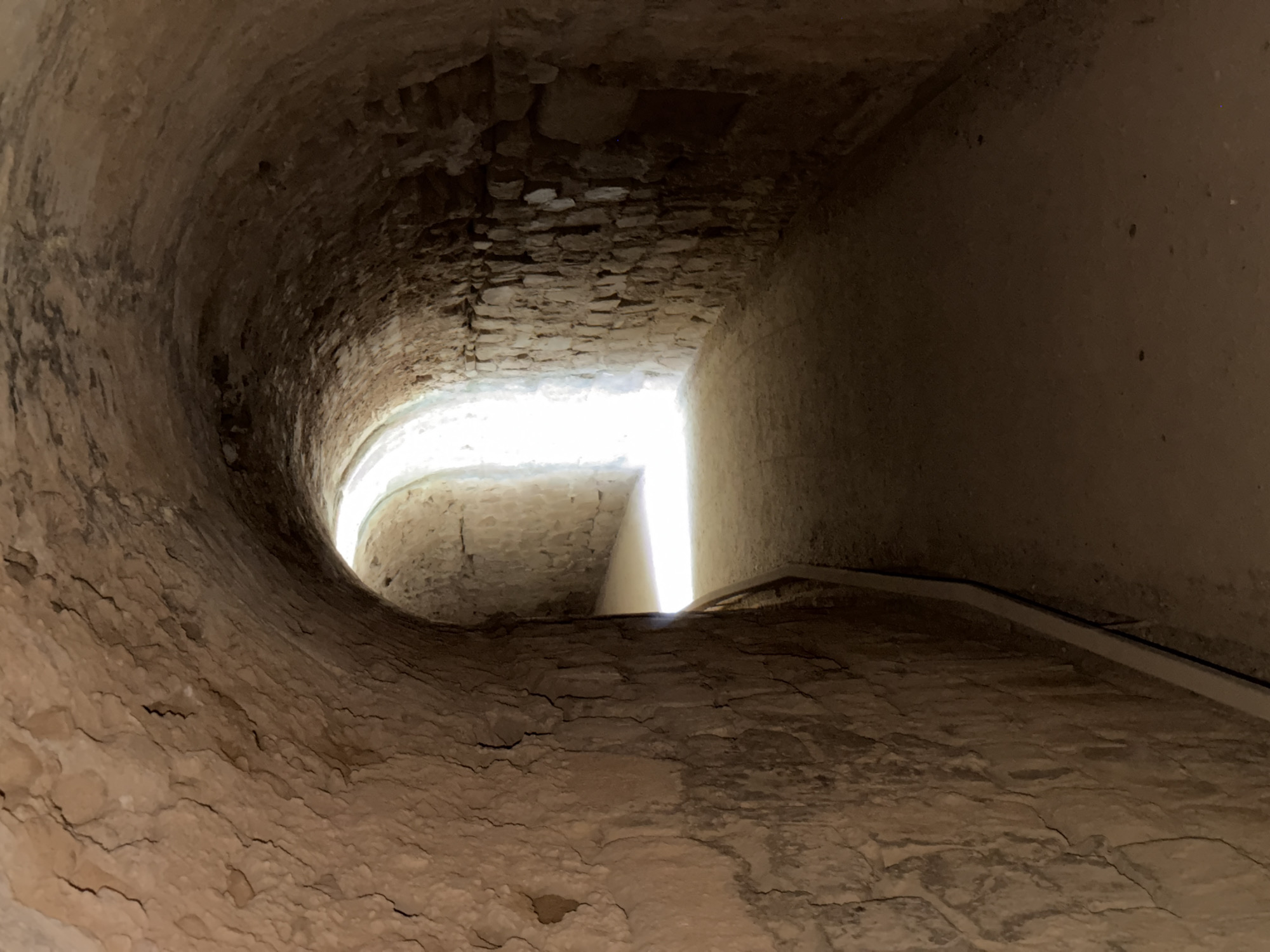 The image shows an ancient stone tunnel. The tunnel is dimly lit, with a bright light source at the end of the tunnel, suggesting an exit or opening. The walls of the tunnel are constructed from rough-hewn stones, with some sections appearing to be made of packed earth or mortar. The floor of the tunnel is covered in dirt and sand. The tunnel appears to be part of an underground structure, possibly an aqueduct or part of Roman ruins. This tunnel is located in Merida, Spain, which is known for its well-preserved Roman ruins.