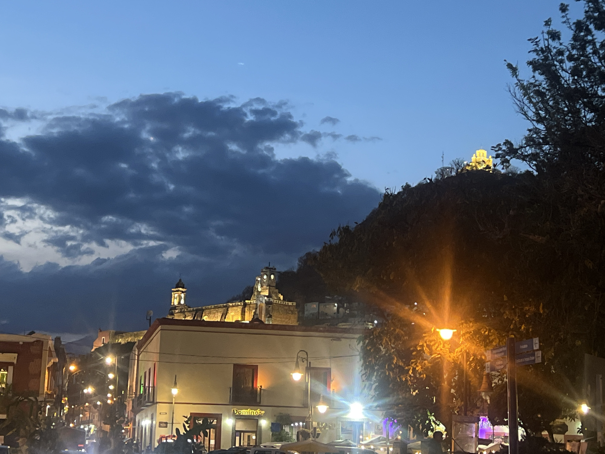 The image depicts a street scene in an urban location at dusk or night. The foreground features a street with illuminated streetlights casting a bright glow. There are buildings on either side of the street. One building has a "Domino's" sign. The buildings appear to be two or three stories high and have lit windows. The presence of parked cars and possibly people under umbrellas suggest an active street scene. In the background, elevated structures, possibly religious buildings or castles, are visible on a hill. The sky is a deep blue with heavy, dark clouds partially covering it. A tree is present on the right side of the frame. The precise location within the city of Unknown, Unknown, cannot be determined without further information.