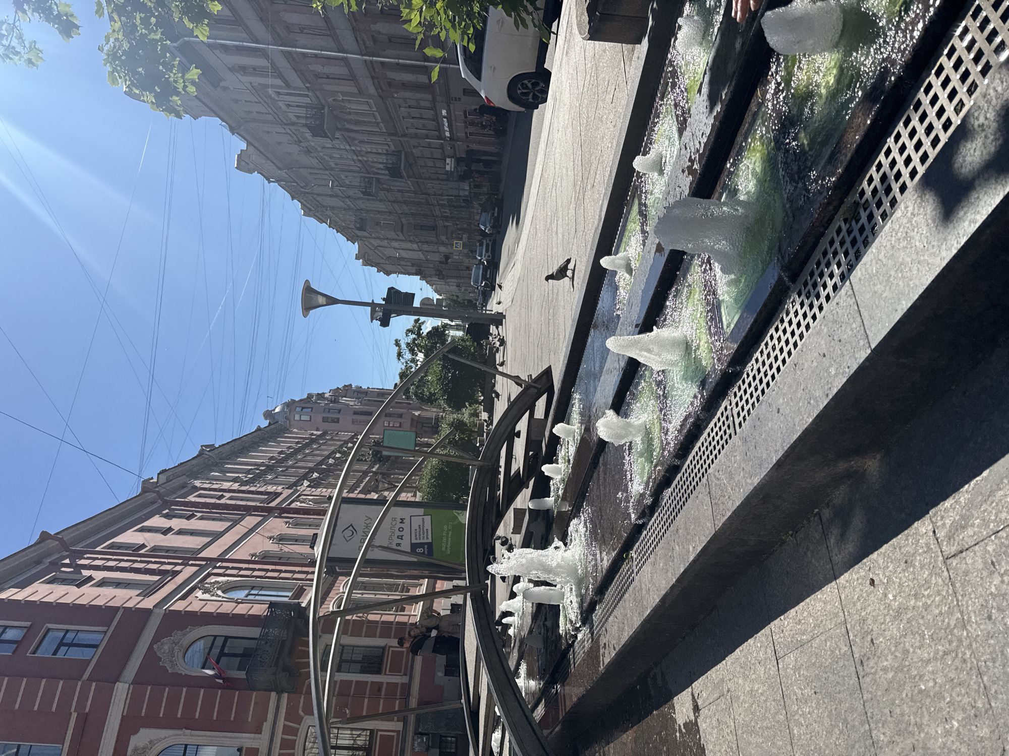 This is a vertical, eye-level shot of a city street scene in Saint Petersburg, Russia, on a bright, sunny day. The image is dominated by the architectural style of the buildings lining the street, with their ornate facades and tall windows. The sky is a clear, brilliant blue, and sunlight streams down, casting distinct shadows.
In the foreground, a contemporary fountain runs along the sidewalk, its jets of water creating a refreshing visual element. The fountain is integrated into the urban landscape, bordered by grey stone paving. A pigeon is visible on the sidewalk near the fountain, adding a touch of everyday life to the scene. Several cars are parked and driving on the street in the middle ground, and a white car is particularly prominent.
The background features more of the historic St. Petersburg architecture, with buildings featuring classical details and repeating window patterns. Overhead, a network of electrical wires crisscrosses the sky, a common sight in many urban environments. A street lamp with a distinctive horn-like shape is visible in the left half of the frame.
There are glimpses of people in the middle and background, mostly indistinct figures on the sidewalk or in cars, suggesting a normal day in the city. The overall mood is one of urban vibrancy under a clear, summer sky. A sign on a metal structure in the middle left of the image contains text in Cyrillic, appearing to advertise a service or event. The specific text is difficult to decipher fully but includes some recognizable Cyrillic letters.
