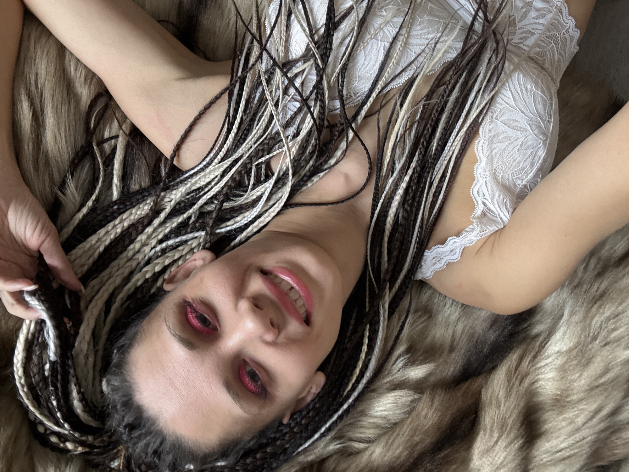 A woman with braided hair lies on a furry surface, possibly a blanket or rug. She is wearing a white lace top and has pink makeup around her eyes and on her lips. She is smiling and holding her hair in her left hand. The background appears to be a neutral color. The image does not provide any information to connect it to Rusanovo, Russia.
