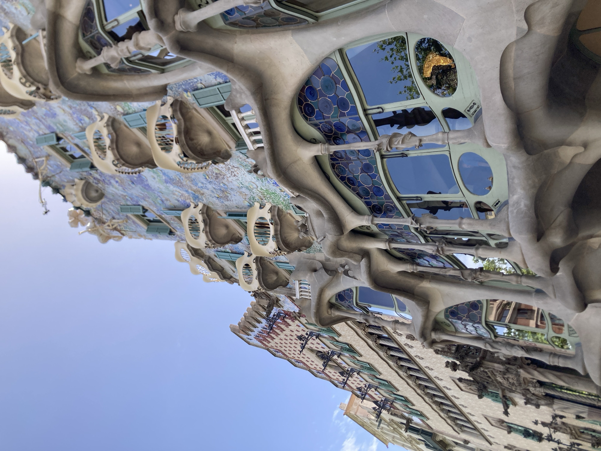The image shows a low-angle, exterior view of the Casa Batlló, a renowned building designed by Antoni Gaudí, located on Passeig de Gràcia in Barcelona, Spain. The facade features a distinctive mosaic of colorful tiles in shades of blue and green, with a wavy, organic design. Several balconies, designed to resemble bone-like structures, project from the building, each with a unique, whimsical shape and adorned with white railings. Green shutters are visible on some windows. The lower part of the building exhibits large, curved windows with intricate stained glass patterns in shades of blue and round, circular motifs. The architectural style is characteristic of Gaudí's Modernisme period, with a focus on natural forms and vibrant colors. The sky above is clear and blue, suggesting a bright, sunny day. The reflection in the stained glass windows shows trees and other buildings, indicating an urban setting.