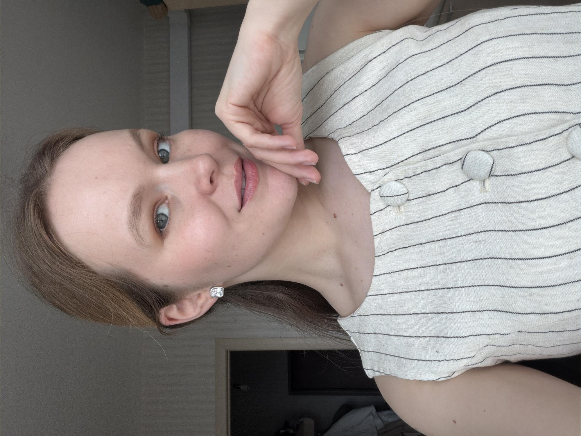A woman is pictured indoors. She has fair skin, light brown hair, and is wearing a striped linen top with button detailing. She is wearing stud earrings and her hand is touching her face. A door can be seen in the background. The location of Izhevsk, Russia is not verifiable.