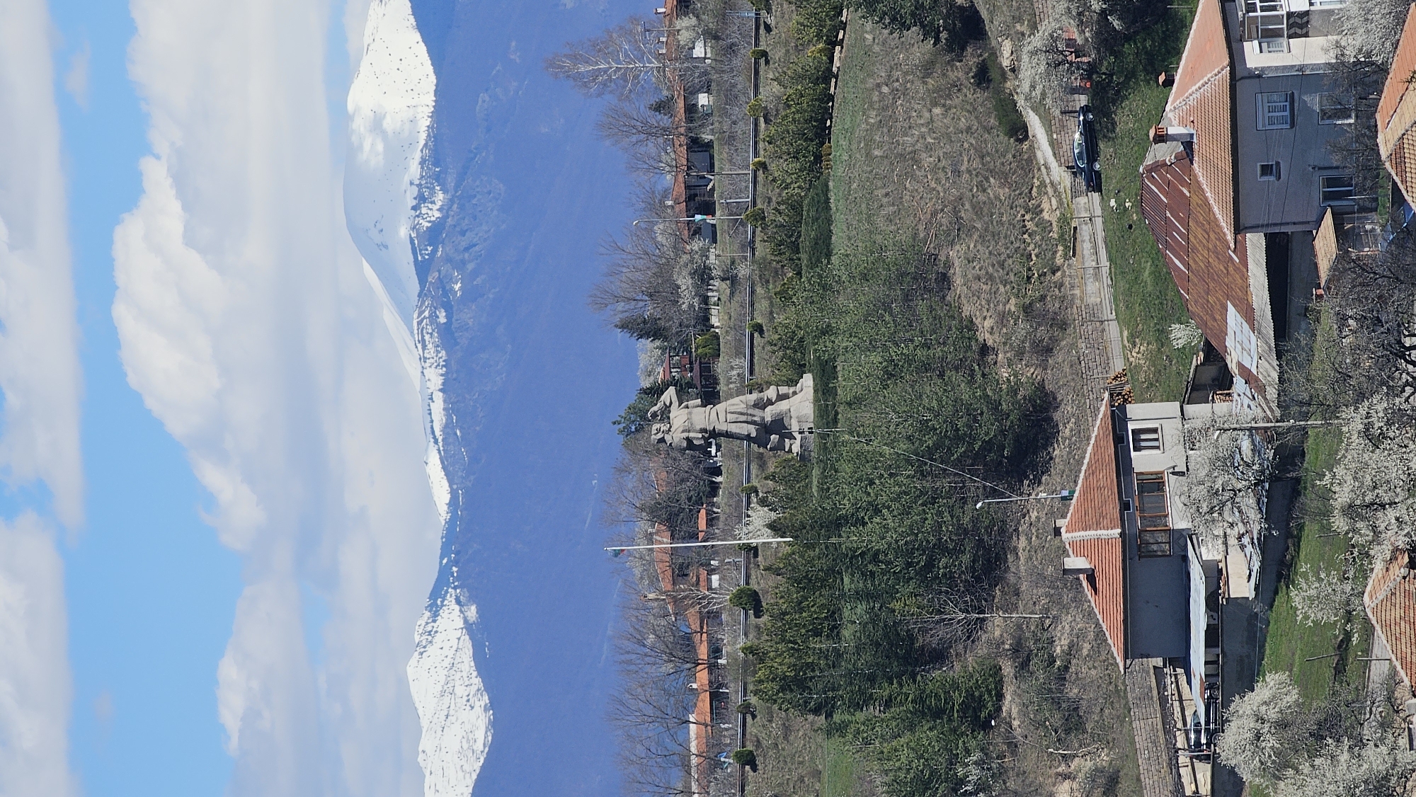 The image shows a view of a landscape with mountains, houses, and trees. On the left side of the image, there is a clear blue sky with some white clouds and a mountain range with snow-covered peaks. In the center of the image, there is a statue of a man on top of a hill, surrounded by trees and vegetation. A road with cars is visible next to the trees, and several houses with orange roofs are on the right side of the image. The landscape appears to be in a town or rural area, with a variety of buildings and natural features. The location is in the city of Unknown, Unknown.
