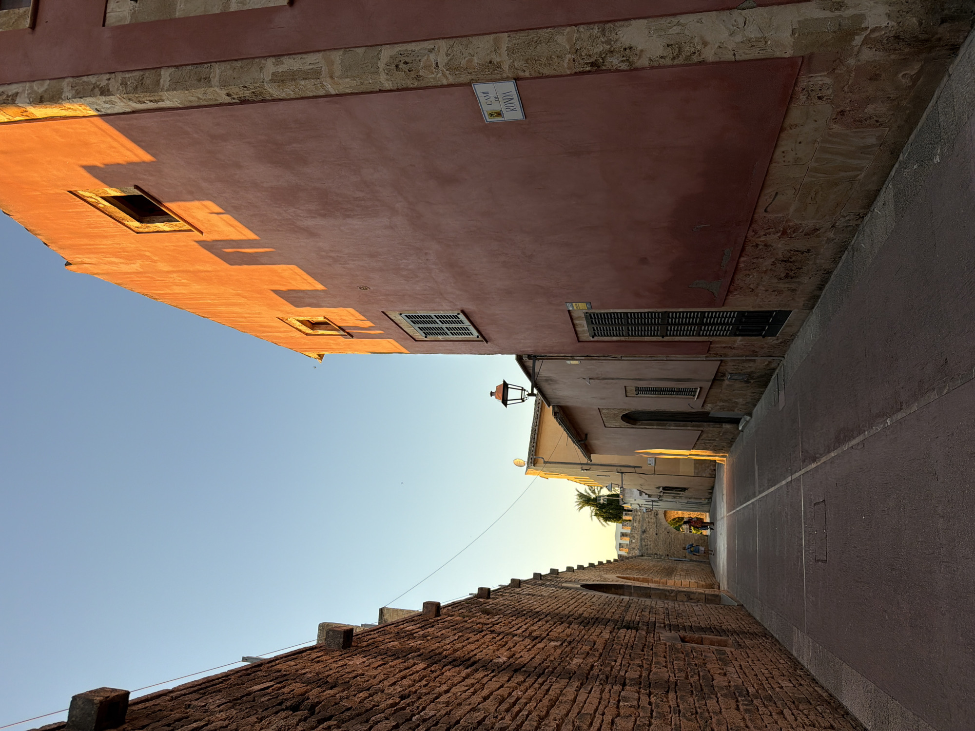 The photograph shows an alleyway in Alcúdia, Spain. On the left is a tall, orange-colored building with small windows casting shadows. A brick wall runs along the bottom left of the image. On the right, another building is visible, with a sign reading "Cami de Ronda." The alleyway is paved and leads towards an archway. Two people, one in blue, are visible in the distance. The sky is a clear blue, fading into a pale yellow where it meets the buildings. A streetlight and some palm trees can be seen near the archway.