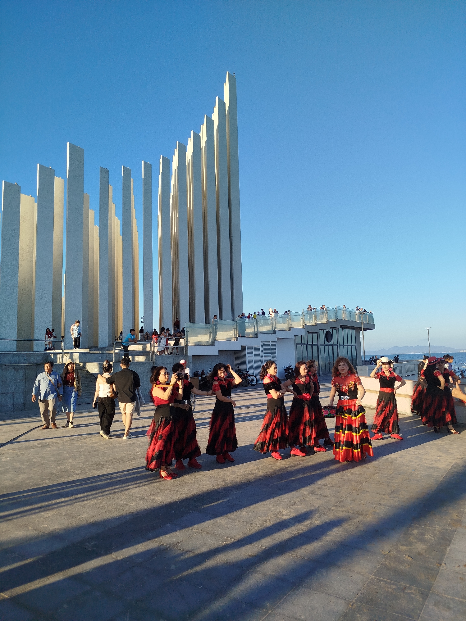 The image depicts a group of women in red and black flamenco-style dresses standing on a concrete plaza. They appear to be posing or preparing for a performance. Behind them is a modern architectural structure composed of tall, white vertical slabs. There are other people walking around the plaza and on an elevated terrace of the building. In the background is a body of water and distant hills. The scene is illuminated by bright sunlight. This appears to be in Ho Chi Minh City, Vietnam.