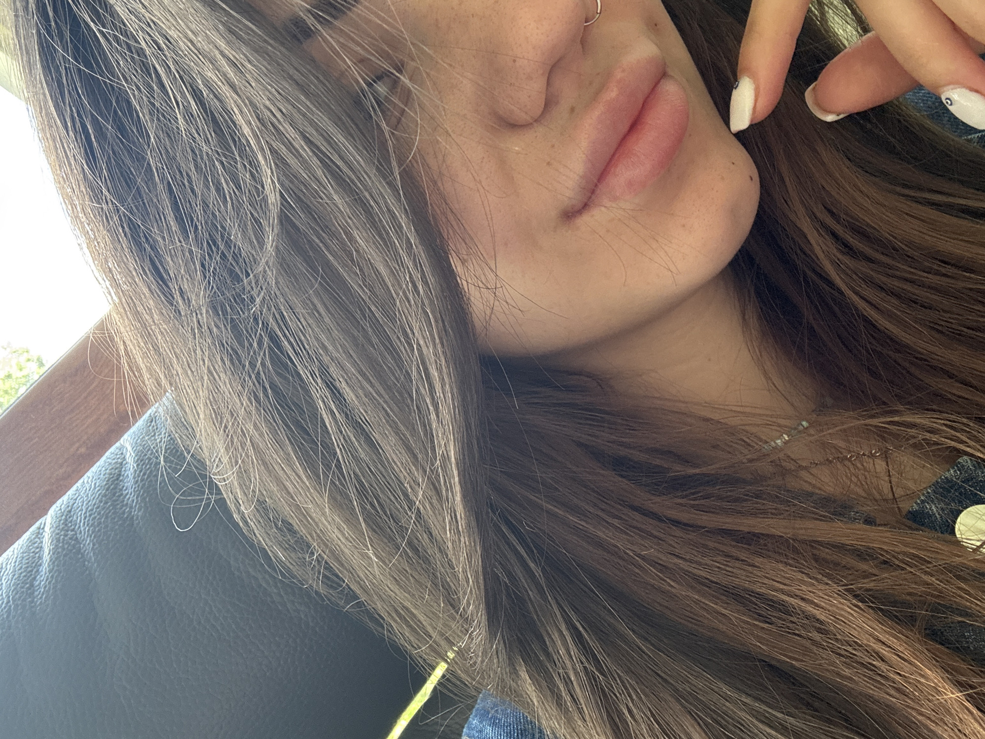 A close-up, angled shot captures the right side of a person's face, primarily focusing on their nose, lips, and chin, with long hair framing the shot. The individual has fair skin with visible freckles across their cheek and nose. A simple, silver-toned hoop nose ring is worn on their right nostril. Their lips are full and appear naturally colored. Long, straight hair, a mix of brown and lighter, possibly ash-blonde or greyish, strands, flows around their face and shoulder. The person's right hand is visible near their chin, with fingers lightly touching their hair. The fingernails are painted white, each featuring a small dark dot near the cuticle. A denim-textured fabric, likely clothing, is visible on the person's right side. In the background, a dark, textured surface, resembling a car seat or couch, is visible, alongside a brightly lit window on the upper left, suggesting an outdoor view during daytime. A thin, silver-toned necklace is subtly visible beneath the hair on the right side of the neck. The scene appears casual, possibly taken inside a vehicle or indoors by a window in Sofia, Bulgaria. No text is visible in the image.