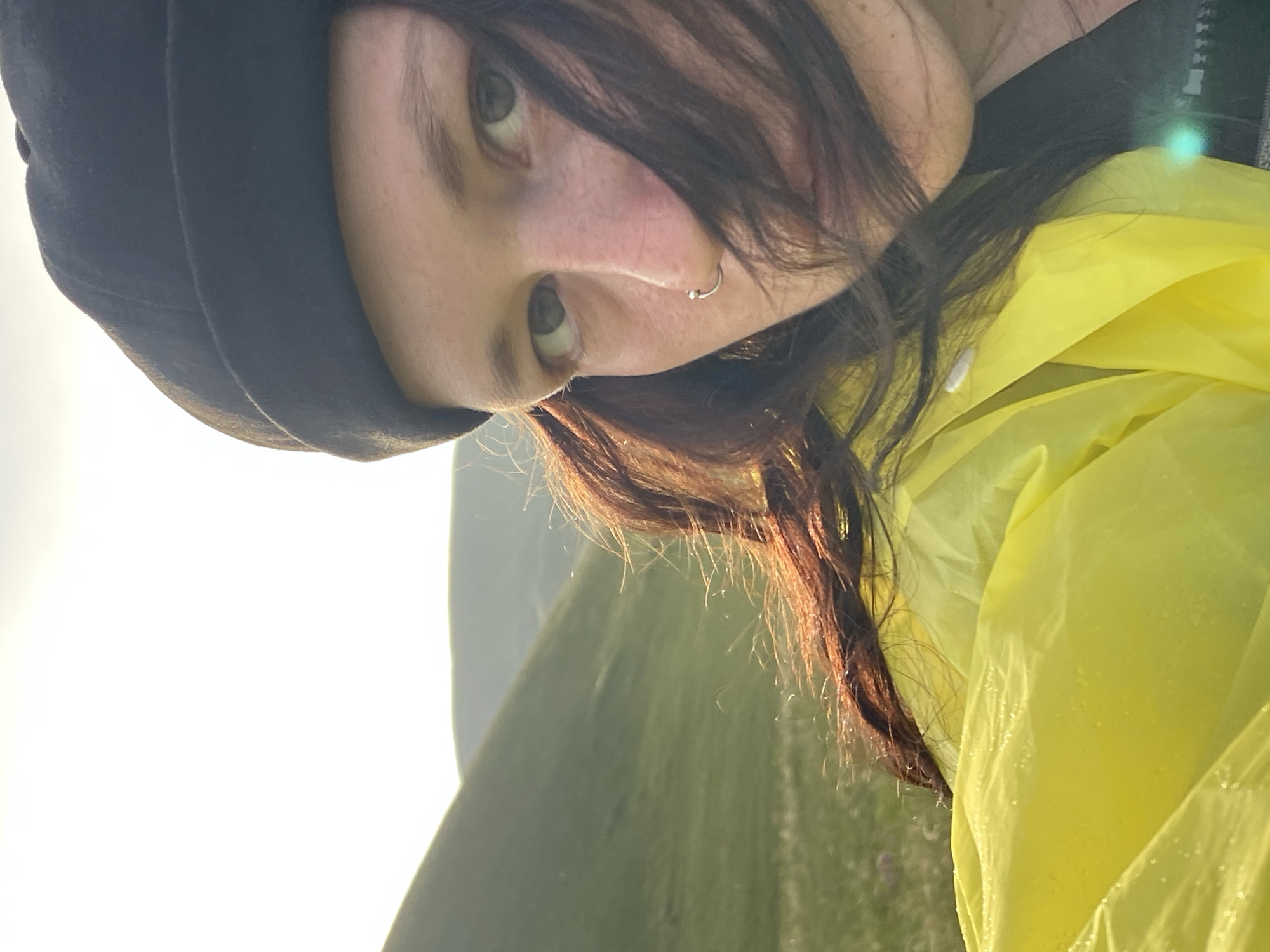 A close-up, selfie-style image shows a person wearing a black beanie and a bright yellow hooded jacket. The person has dark hair that is partially obscuring their face, and a silver nose ring. Their eyes are open and looking towards the camera. The background is a landscape of rolling green hills, suggesting an outdoor, possibly rural or mountainous location. The sky is bright and appears to be either sunrise or sunset, with the sun creating a strong backlight. The image quality is good, with natural lighting and sharp focus on the person's face. There are no visible elements that specifically indicate the location is the city of Donskoe, Russia.
